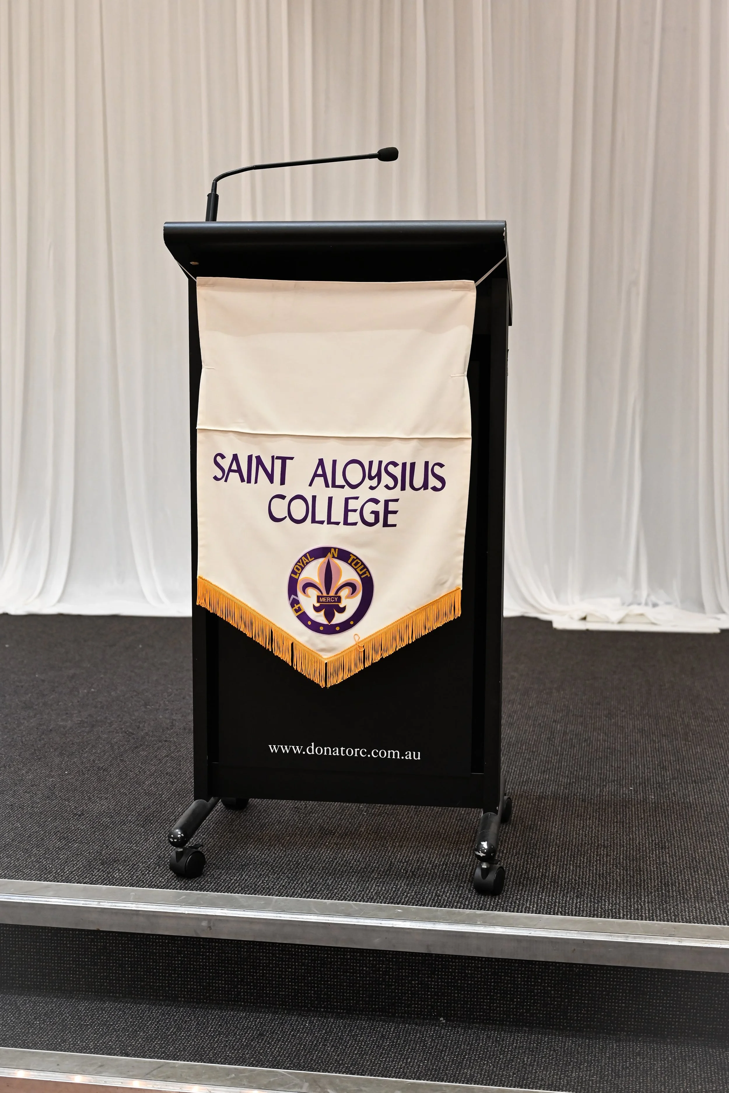 A black podium with a beige banner hanging on the front. The banner displays the name 'Saint Aloysius College' and an emblem with a fleur-de-lis, and the words 'Loyal', 'N', 'Tout', and 'Mercy' inside a circle. The podium has a microphone attached at
