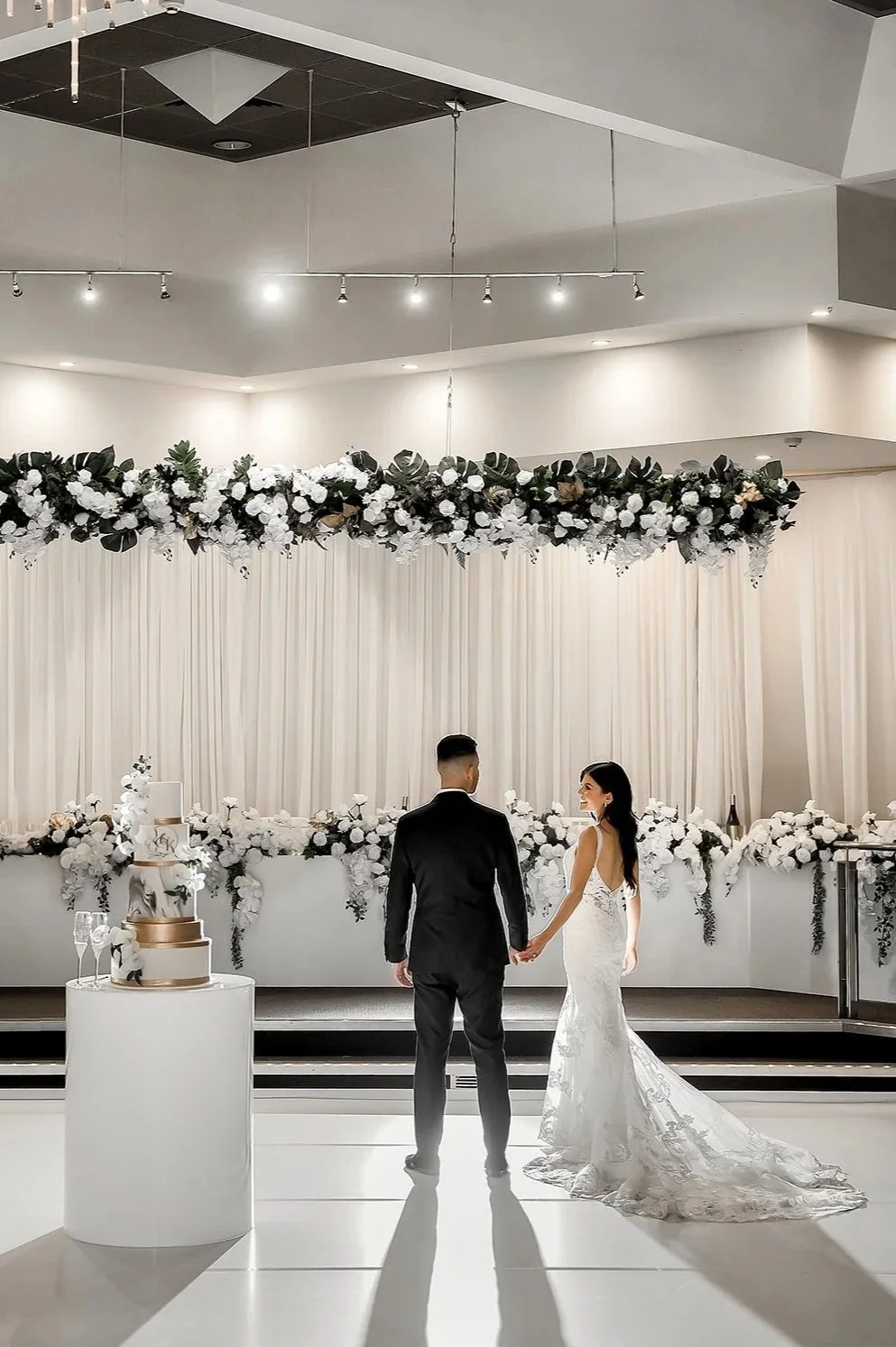 A bride and groom holding hands on a dance floor during their wedding reception, with a decorated backdrop of white and dark flowers and a wedding cake in the foreground at Donato reception centre in Adelaide