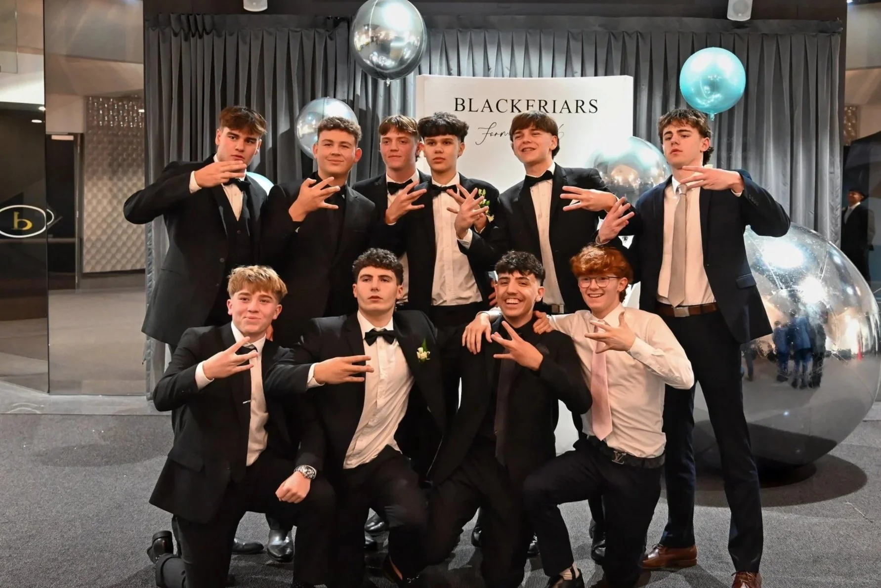 Group of young men in formal suits and ties posing for photo at event, with 'Blackfriars' sign and decorative balloons in background.