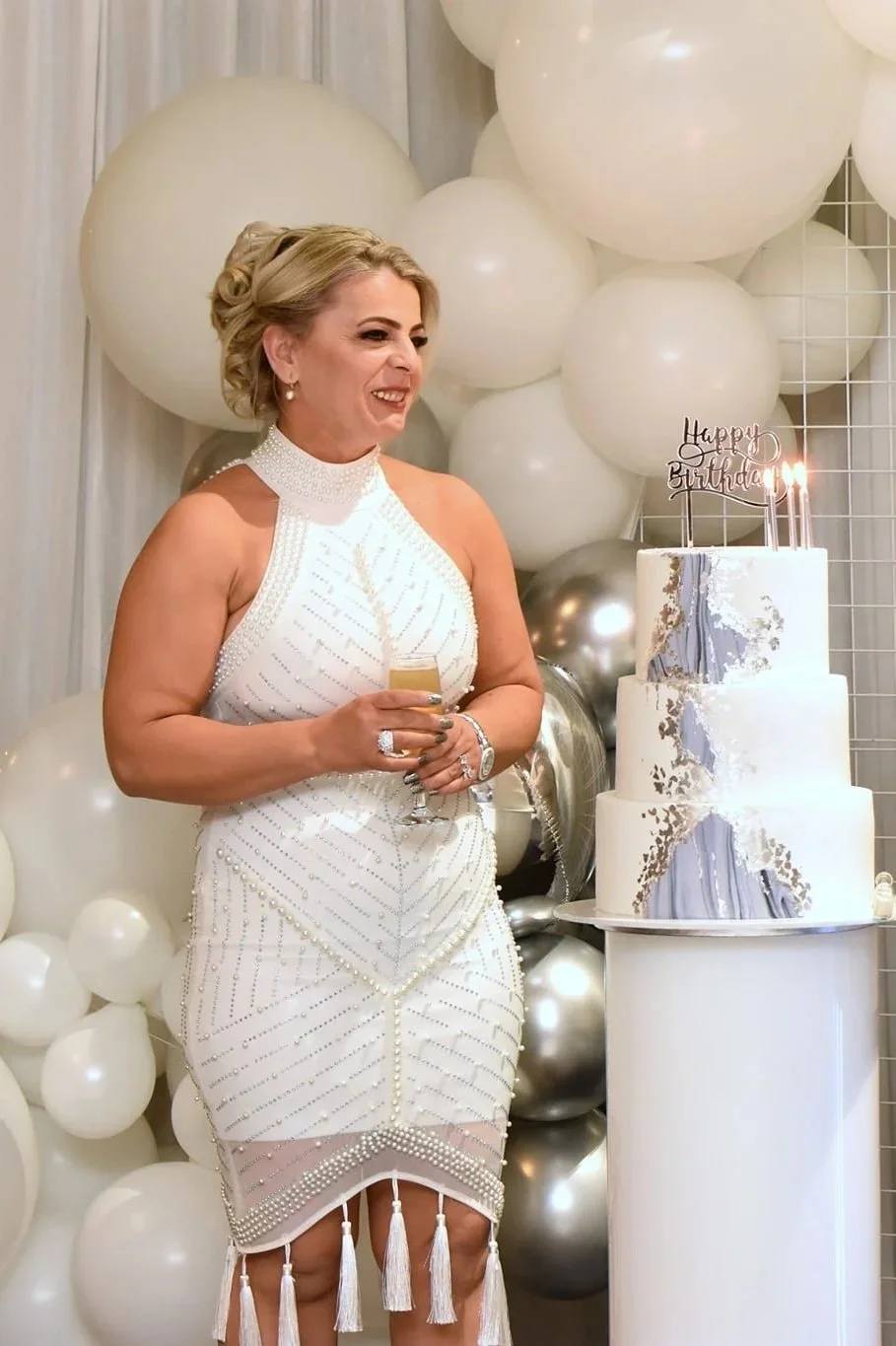 A woman wearing a white, beaded dress standing near a birthday cake with lit candles, surrounded by white and silver balloons, holding a glass of champagne smiling.