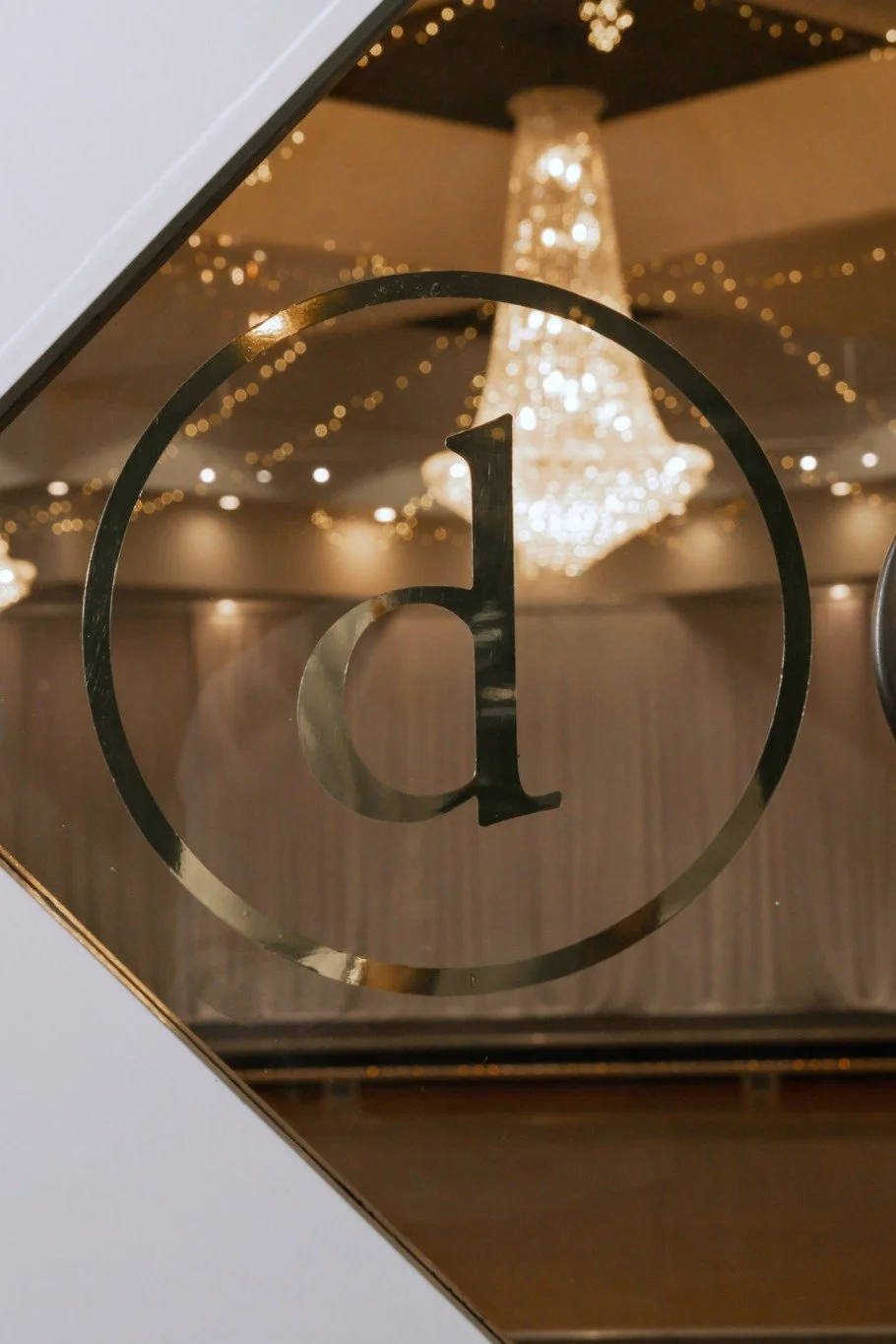 Close-up of a glass panel with a black lowercase letter 'd' inside a circle, bugle-shaped chandelier and string lights reflected in the background.