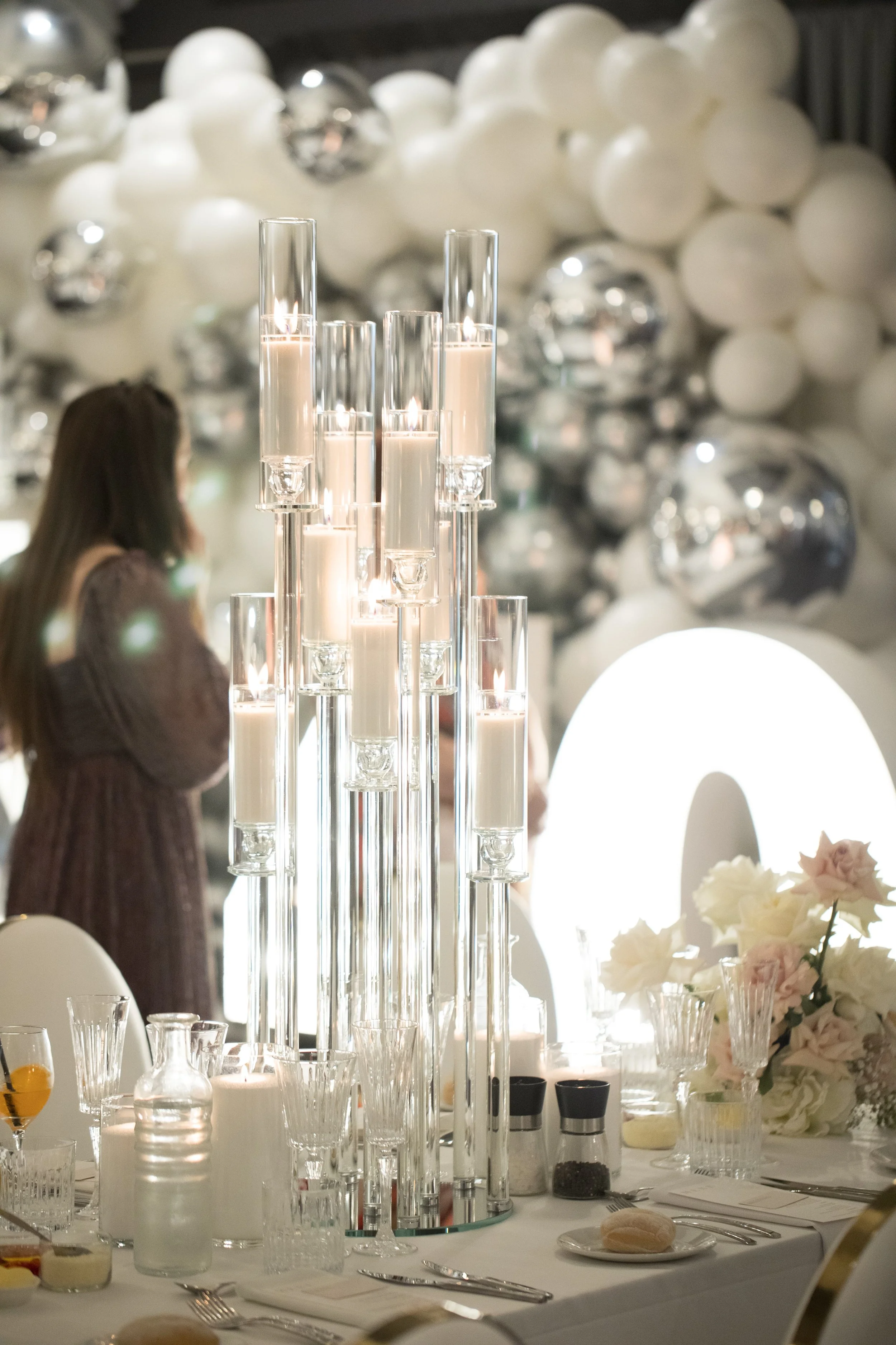 Elegant banquet table setup with tall glass candle holders, white candles, wine glasses, water bottles, salt and pepper shakers, a bread roll on a plate, and floral arrangements, with white balloons and shiny silver ornaments in the background.