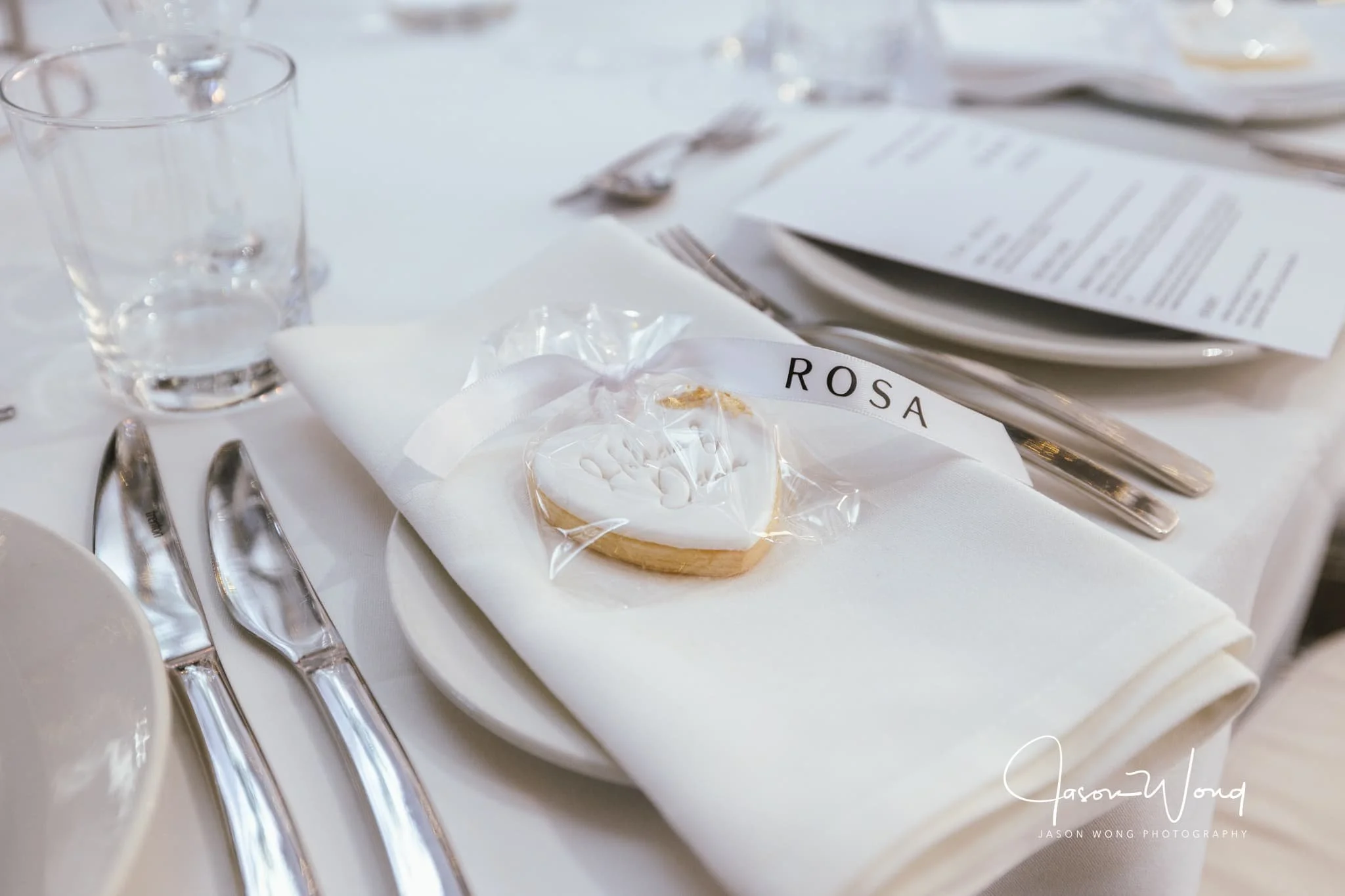 Table setting with glassware, silverware, a white napkin, and a small decorated cookie labeled 'Rosa' at a formal event.