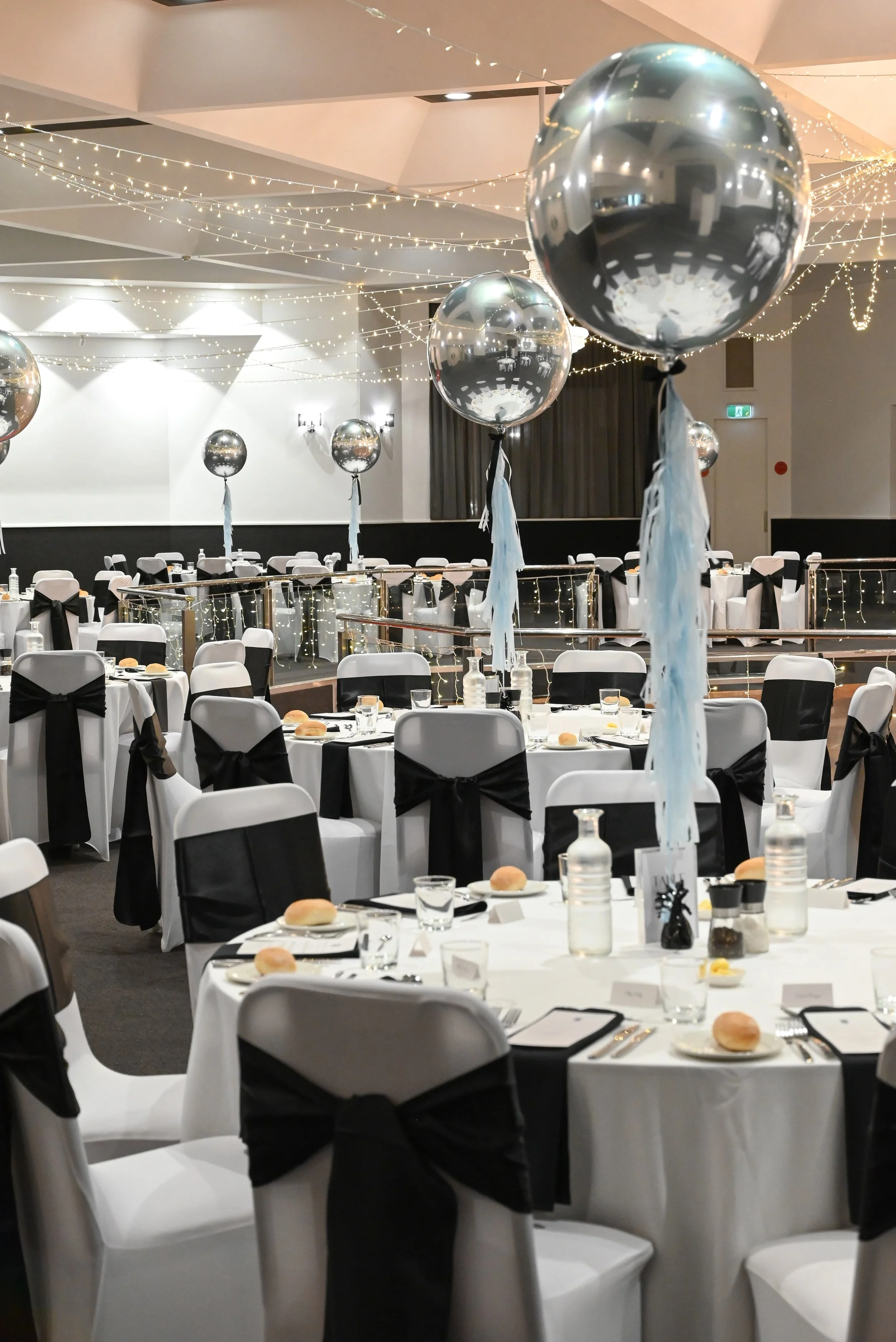 Decorated banquet hall with tables set for an event, black and white color theme, large silver balloons with white ribbons, string lights hanging from the ceiling.