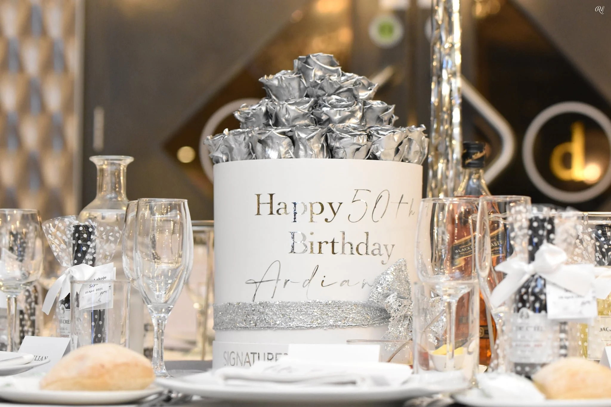 A birthday celebration table with a white cake decorated with silver wrapping paper and the message "Happy 50th Birthday Adrian" written on it. The table includes champagne glasses, bottles, and party decorations in a festive setting.
