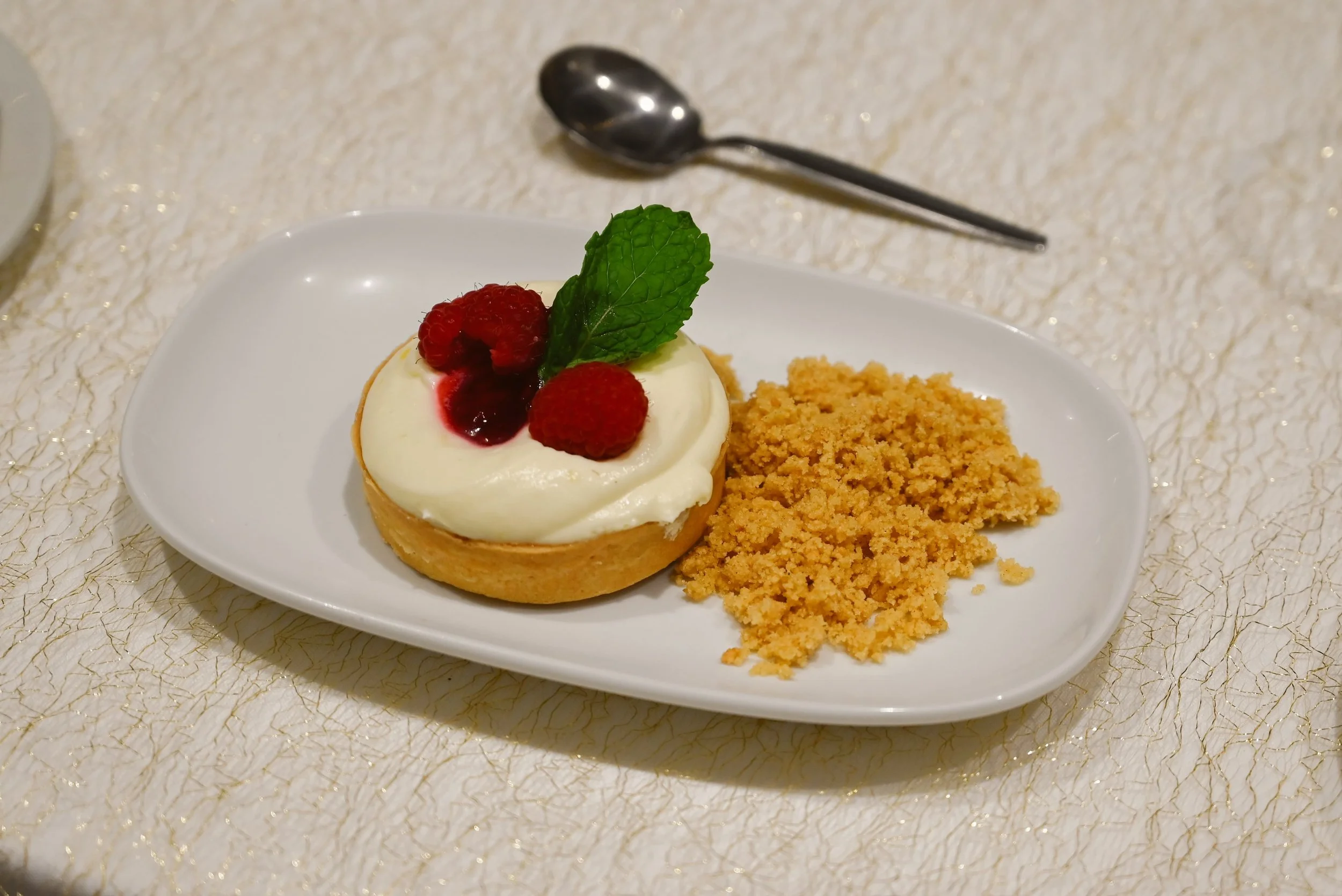 A dessert plate with a cheesecake topped with raspberries, a mint leaf, and raspberry sauce, alongside a serving of crumbled cookie or graham cracker crust. There is a silver spoon in the background on a textured, gold-accented tablecloth.