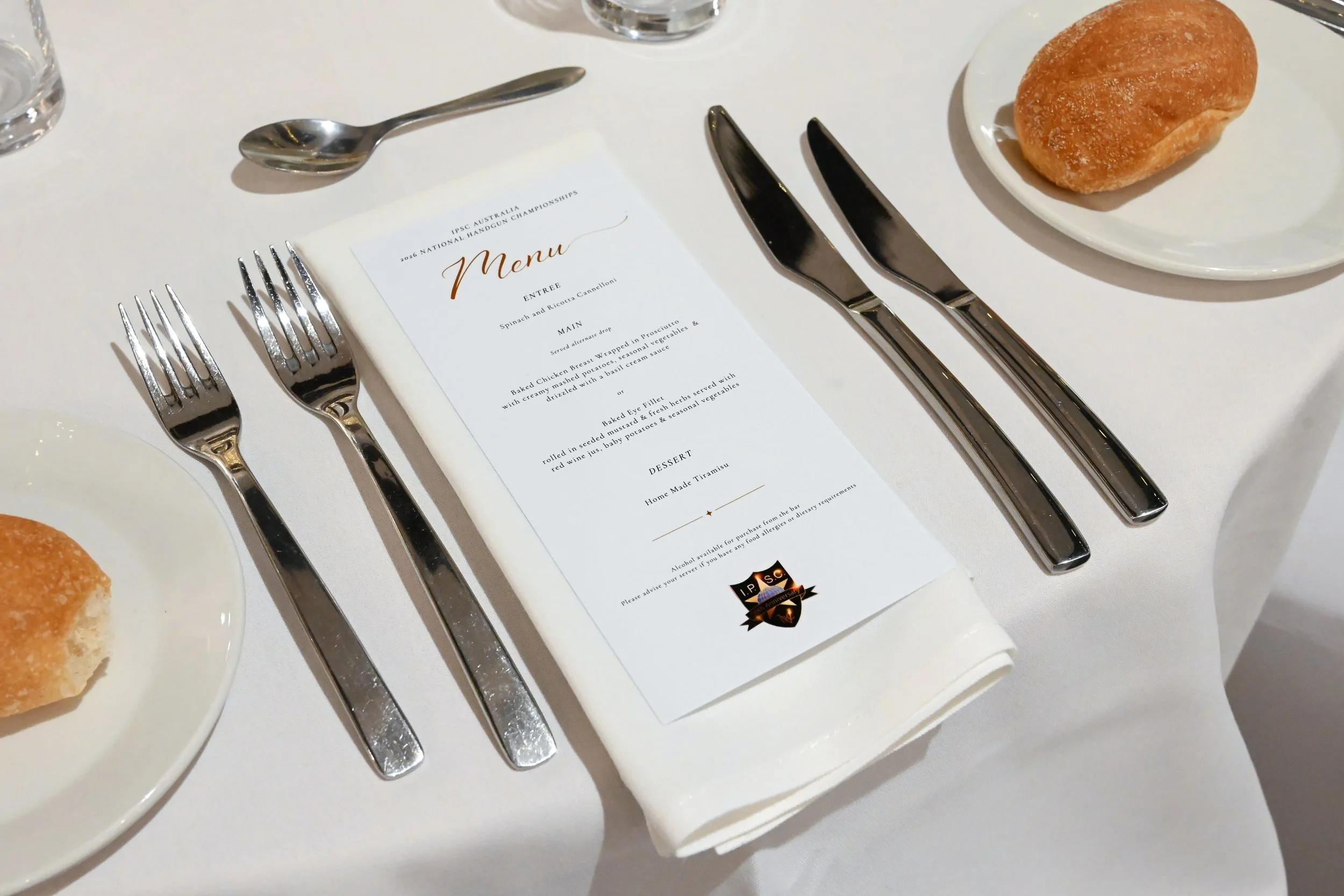 A table setting with a menu, silverware, a bread roll on a plate, and a glass of water.