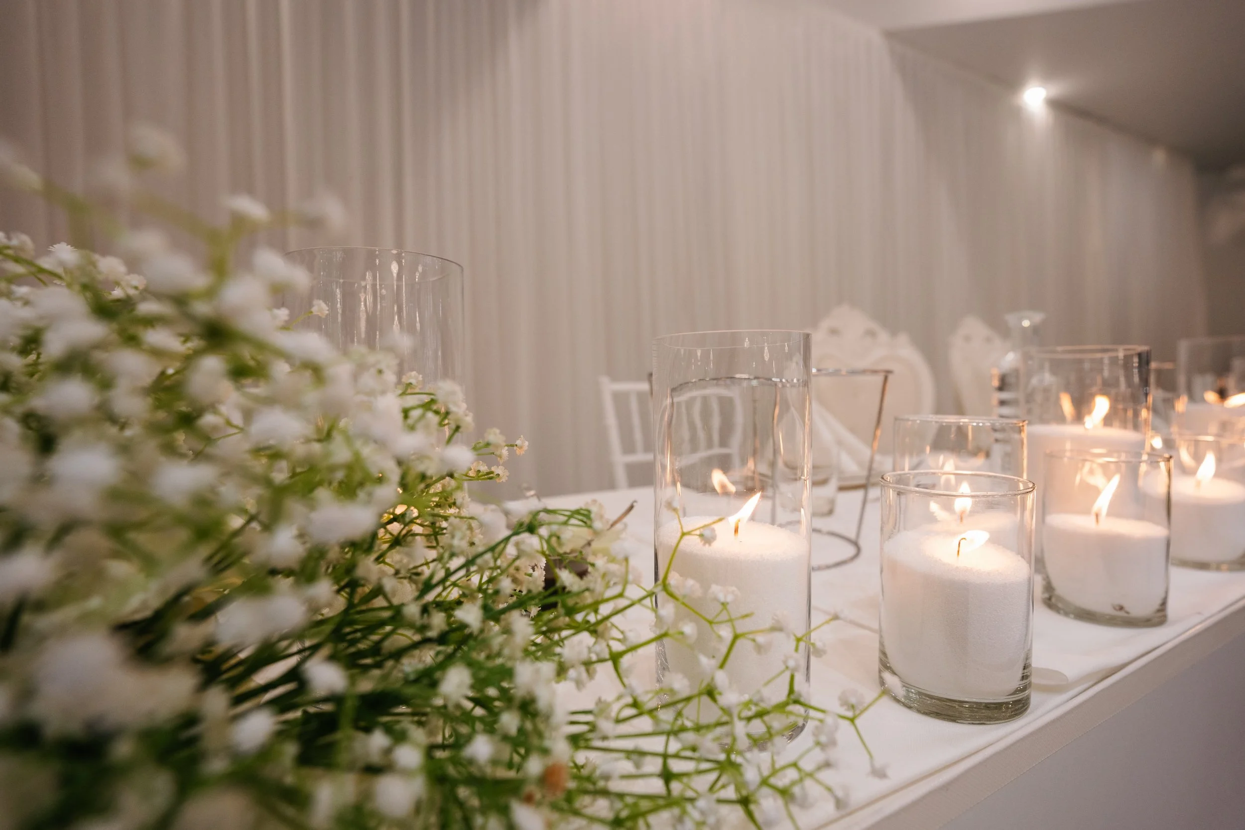 Elegant table setting with white flowers and candles in glass holders on a white table, against a beige and white striped wall.