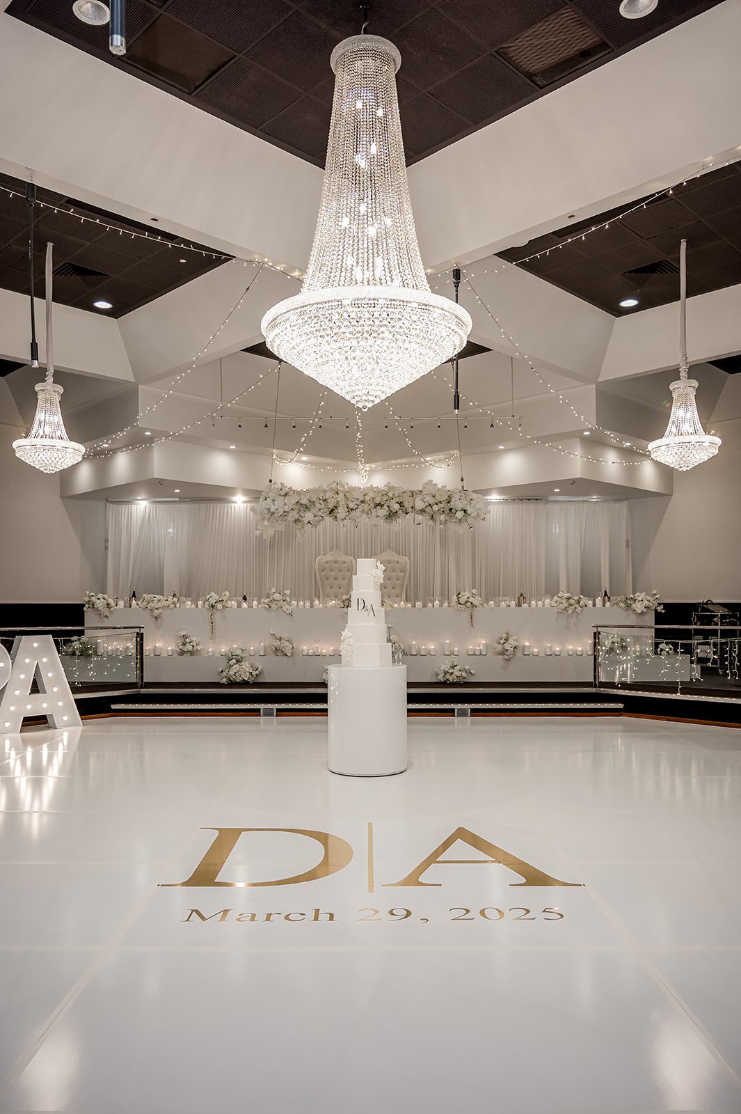Elegant wedding reception hall decorated in white with a large crystal chandelier, floral arrangements, candles, and a tiered wedding cake on a white pedestal at Donato reception centre in Adelaide