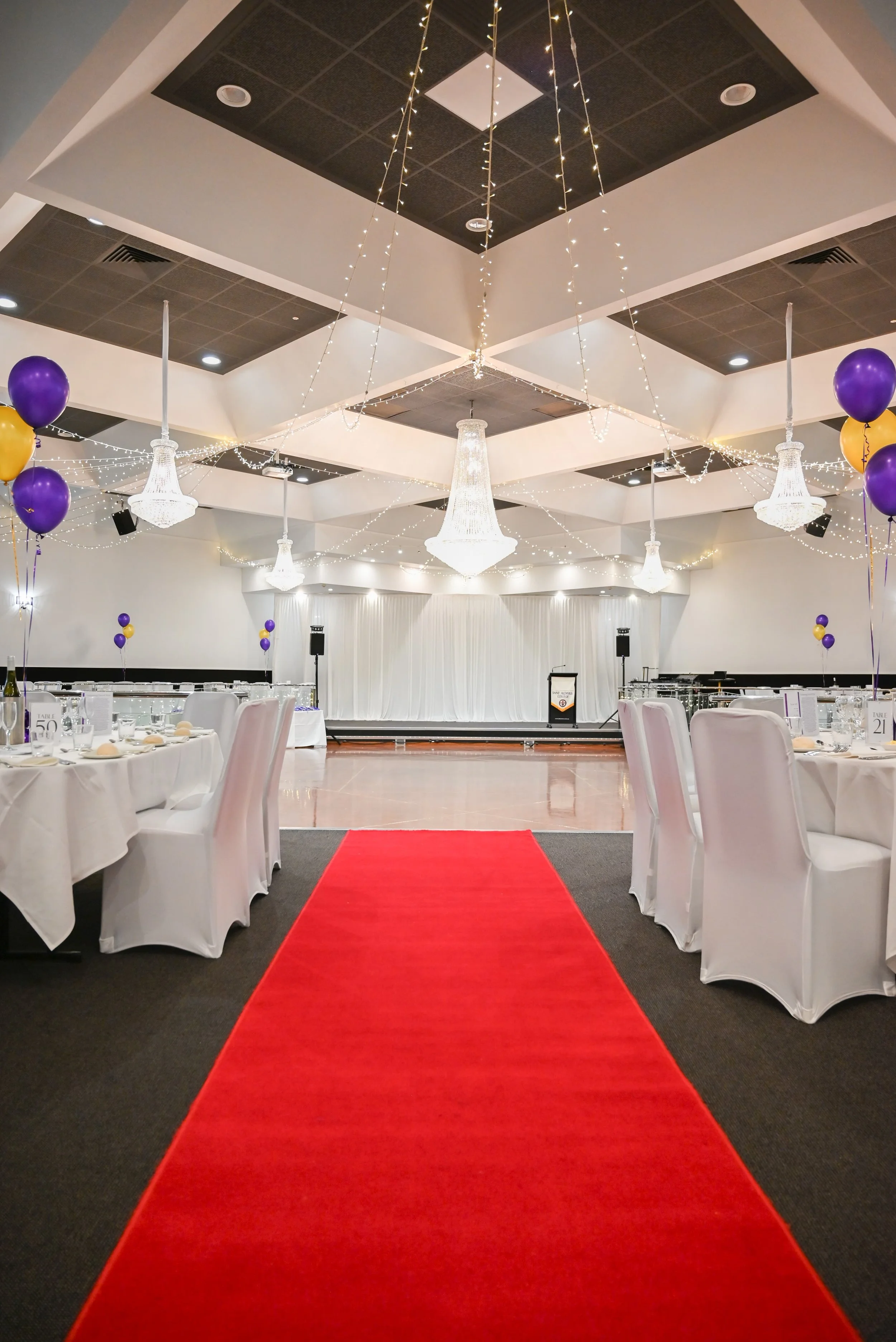 Empty banquet hall decorated for an event with purple and gold balloons, white tablecloths, folded napkins, chandeliers, fairy lights, a red carpet aisle, and a stage at the back.
