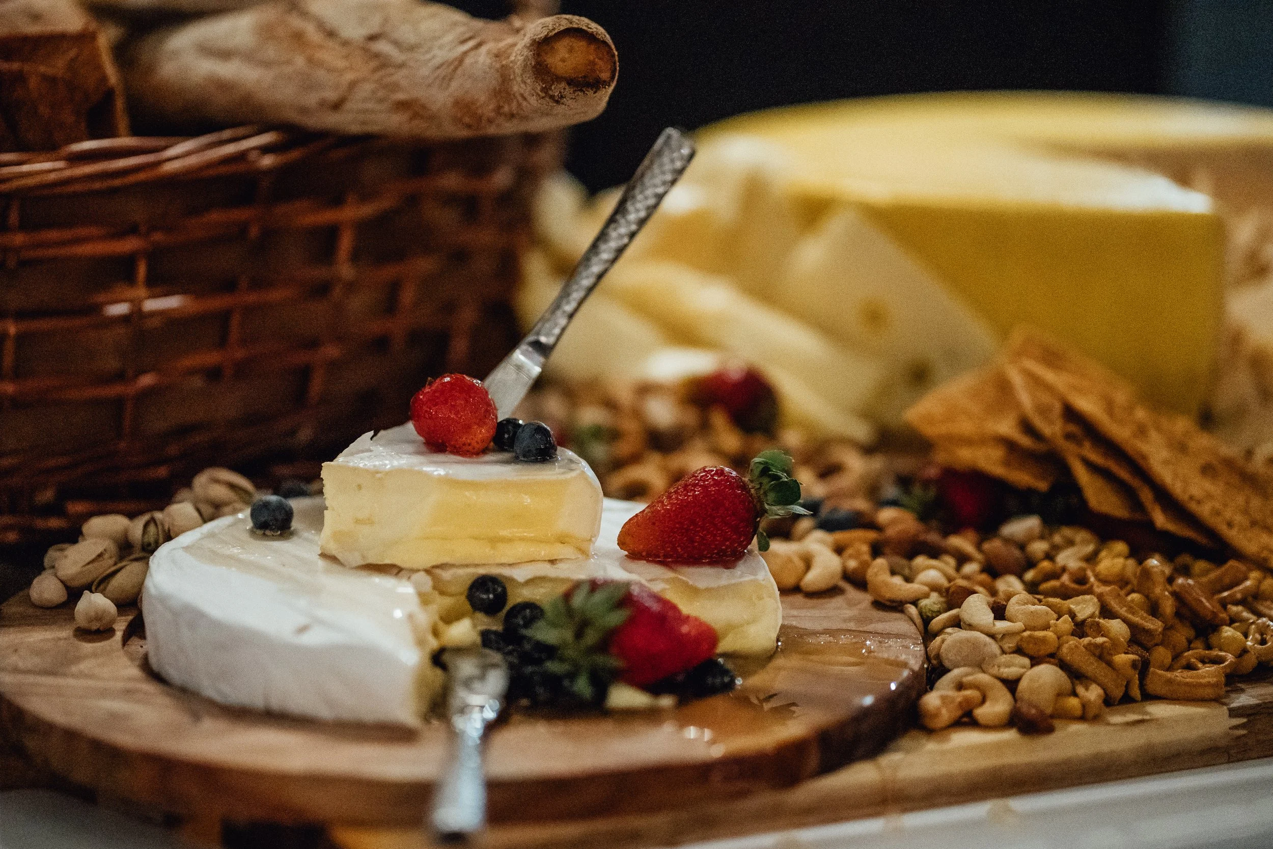 Close-up of a cheese wheel with a piece cut out, topped with strawberries and blueberries, on a wooden board surrounded by nuts and dried fruit. In the background, there is a wicker basket, cinnamon sticks, and a block of yellow cheese.