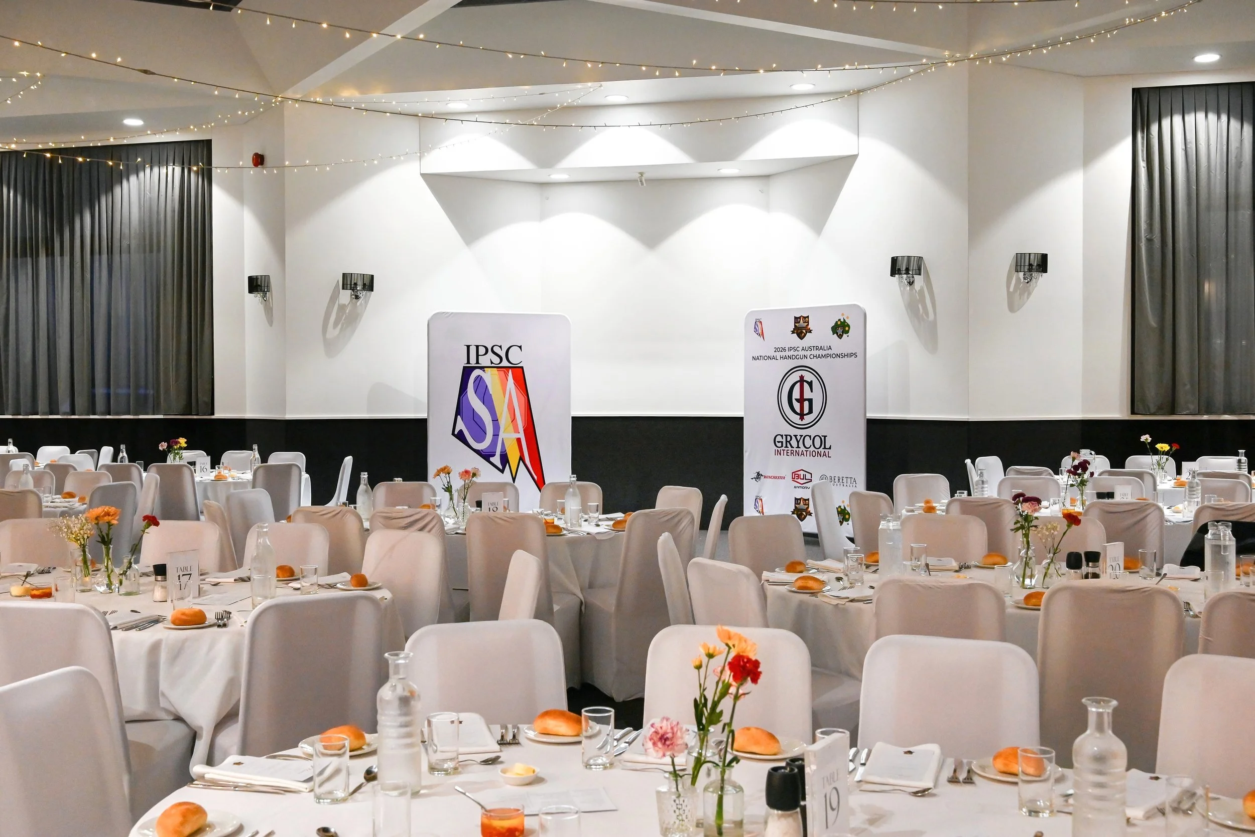 Banquet hall with round tables set for an event, decorated with flowers and bread rolls, with a large stage backdrop featuring the logos of IPSC and Grycol International. String lights adorn the ceiling.