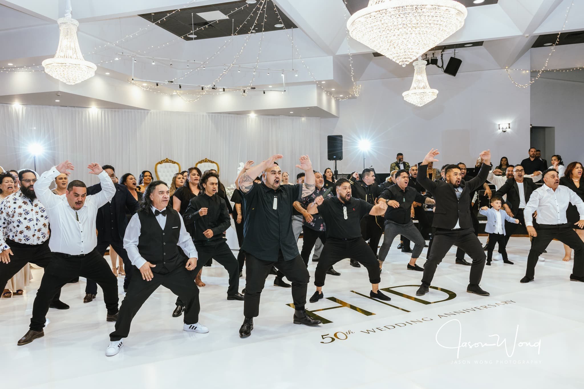 Group of people dancing and singing during a celebration in a decorated hall with chandeliers and fairy lights. The floor has gold accents, and a sign reads '50th Wedding Anniversary'.