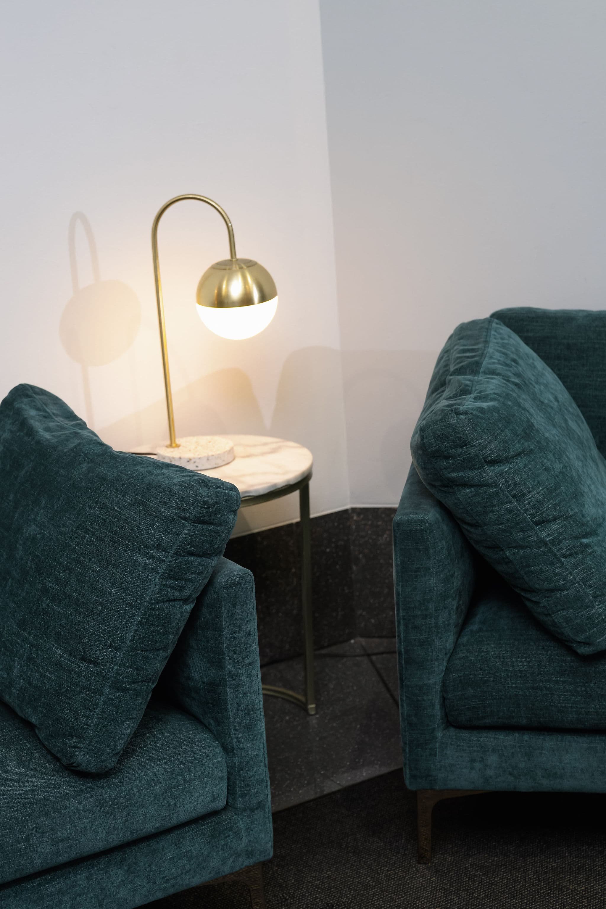 Close-up of two teal upholstered armchairs with matching cushions in a living room, facing toward a small round side table with a marble top. On the table, there is a brass and glass table lamp casting a warm light.