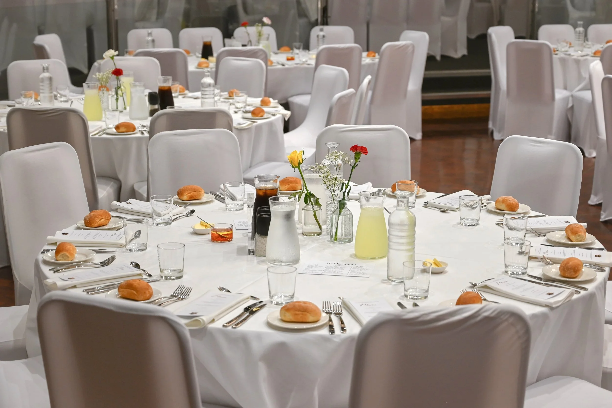 Wedding reception table set with white tablecloths, white chairs, and centerpieces of flowers in vases. The table is decorated with bread rolls, water bottles, glasses, and menus.