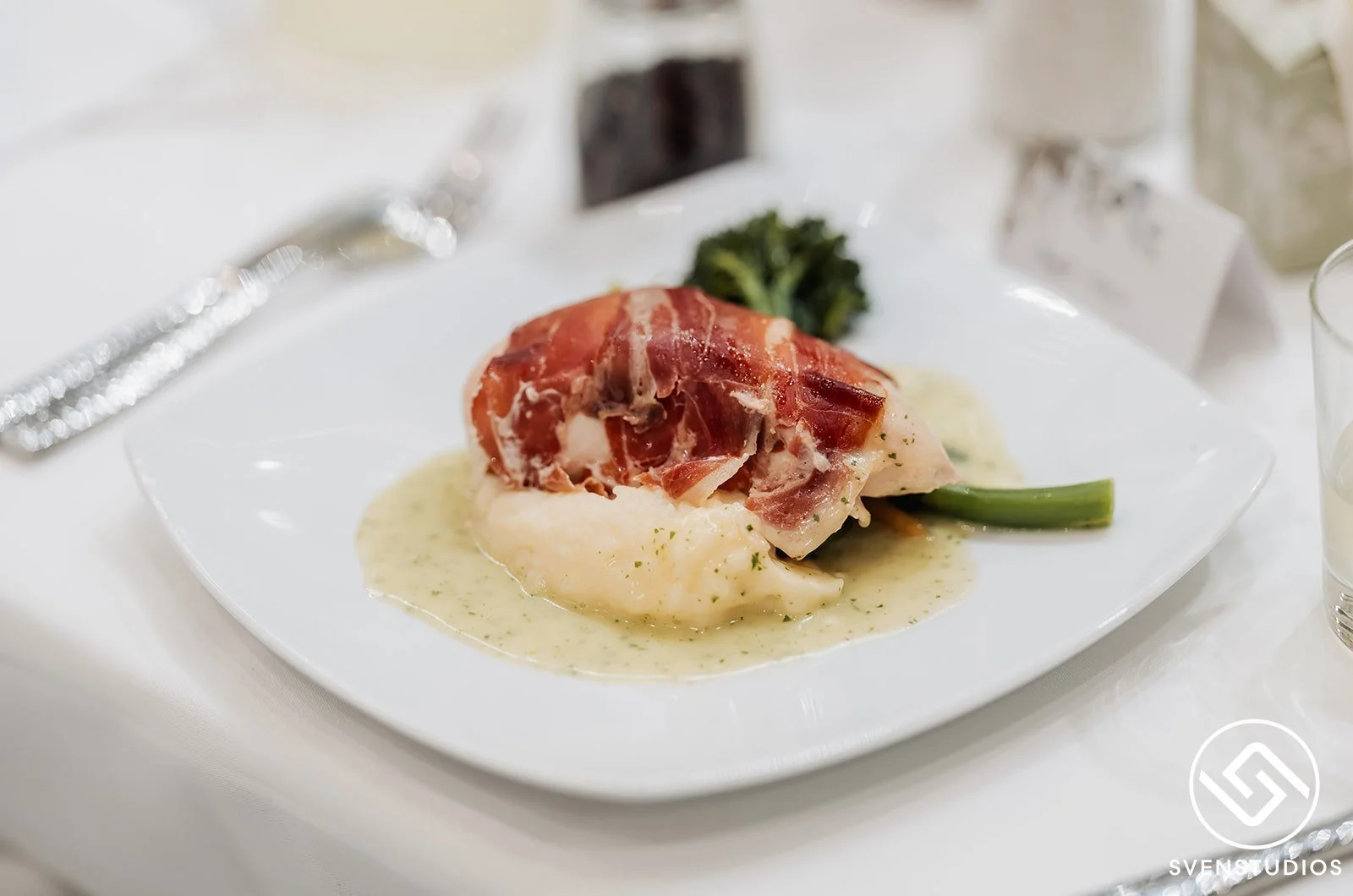 Plate of food with a piece of fish topped with prosciutto, served with a creamy sauce, green beans, and broccoli in the background.
