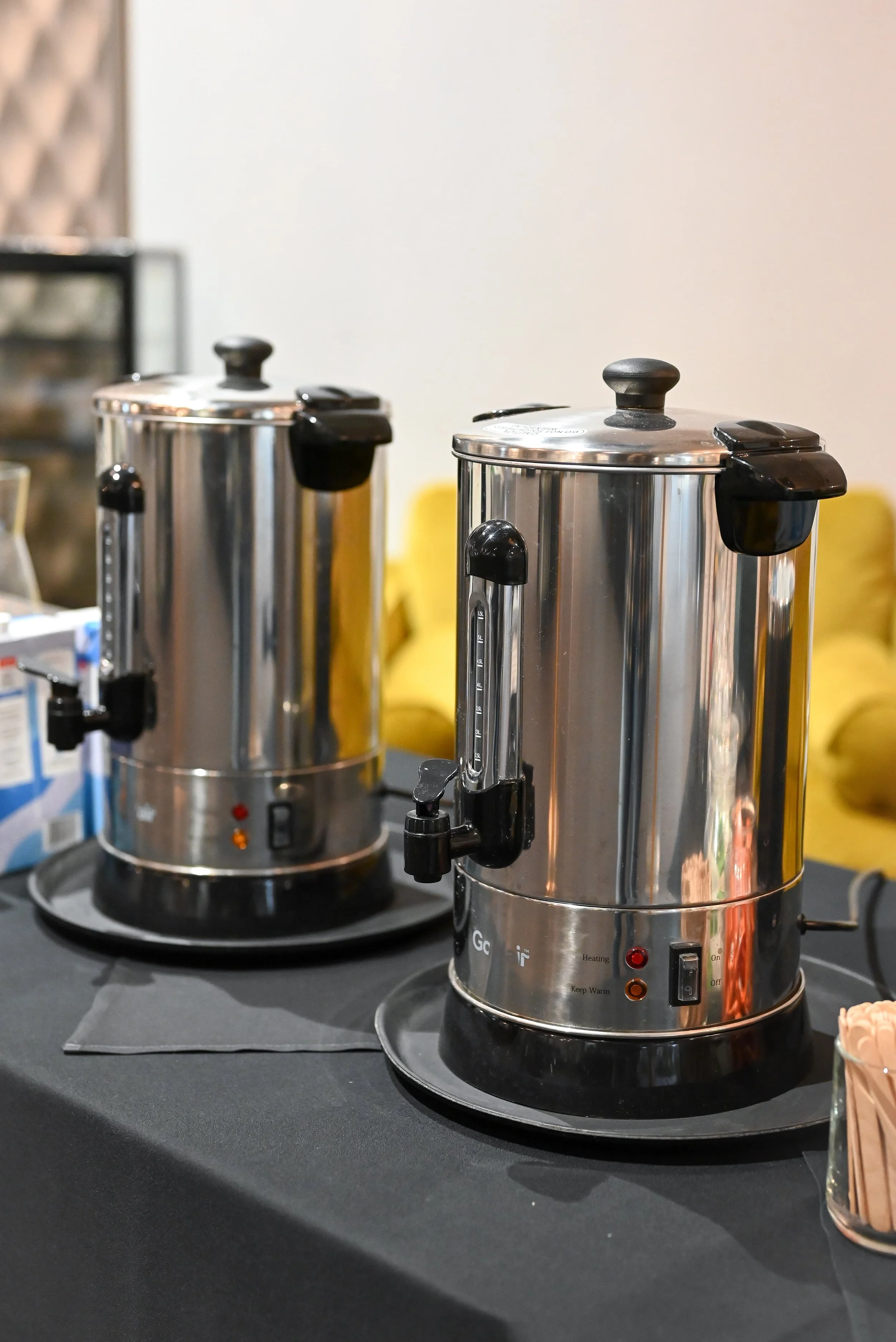 Two stainless steel hot beverage dispensers on a black countertop, with control buttons and red indicator lights, set in an indoor environment.