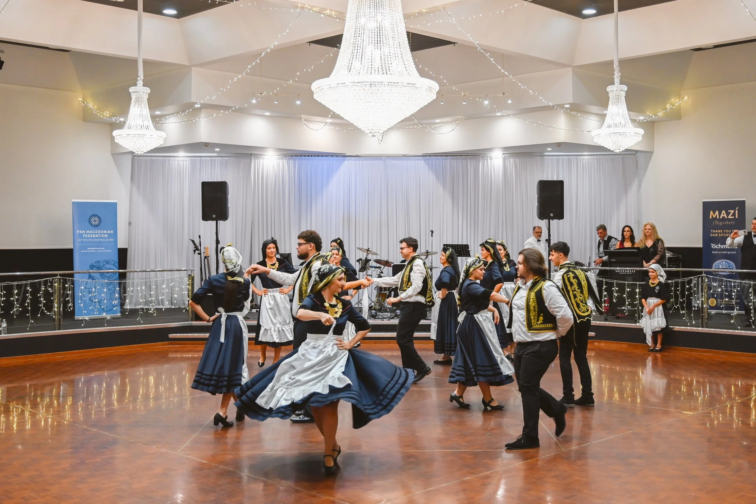 A group of people dressed in traditional Central European clothing, dancing in a ballroom decorated with chandeliers and string lights, with musical instruments on stage in the background.