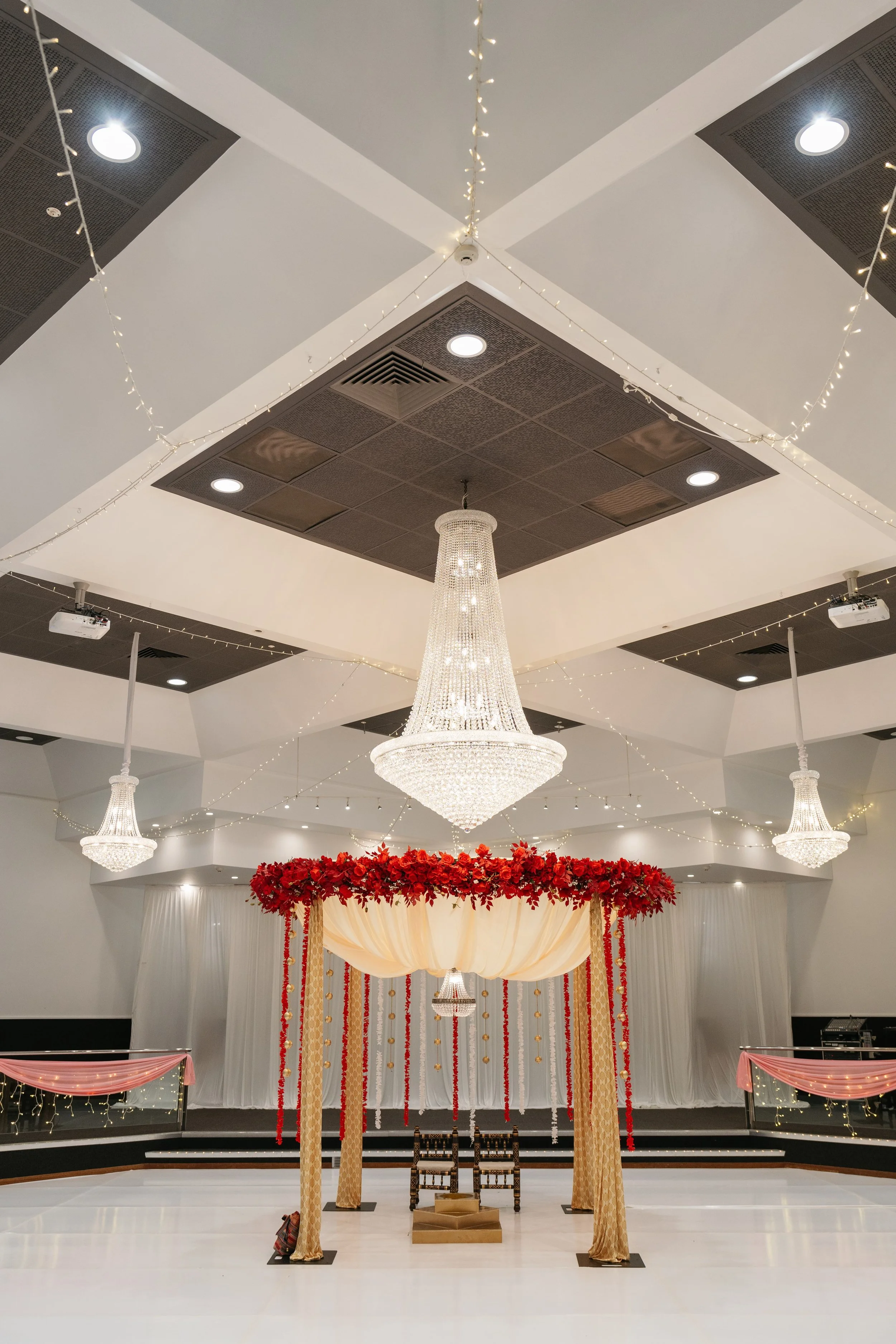Wedding stage decorated with red roses and draped curtains, with chandeliers hanging from the ceiling, and an ornate backdrop for the ceremony.