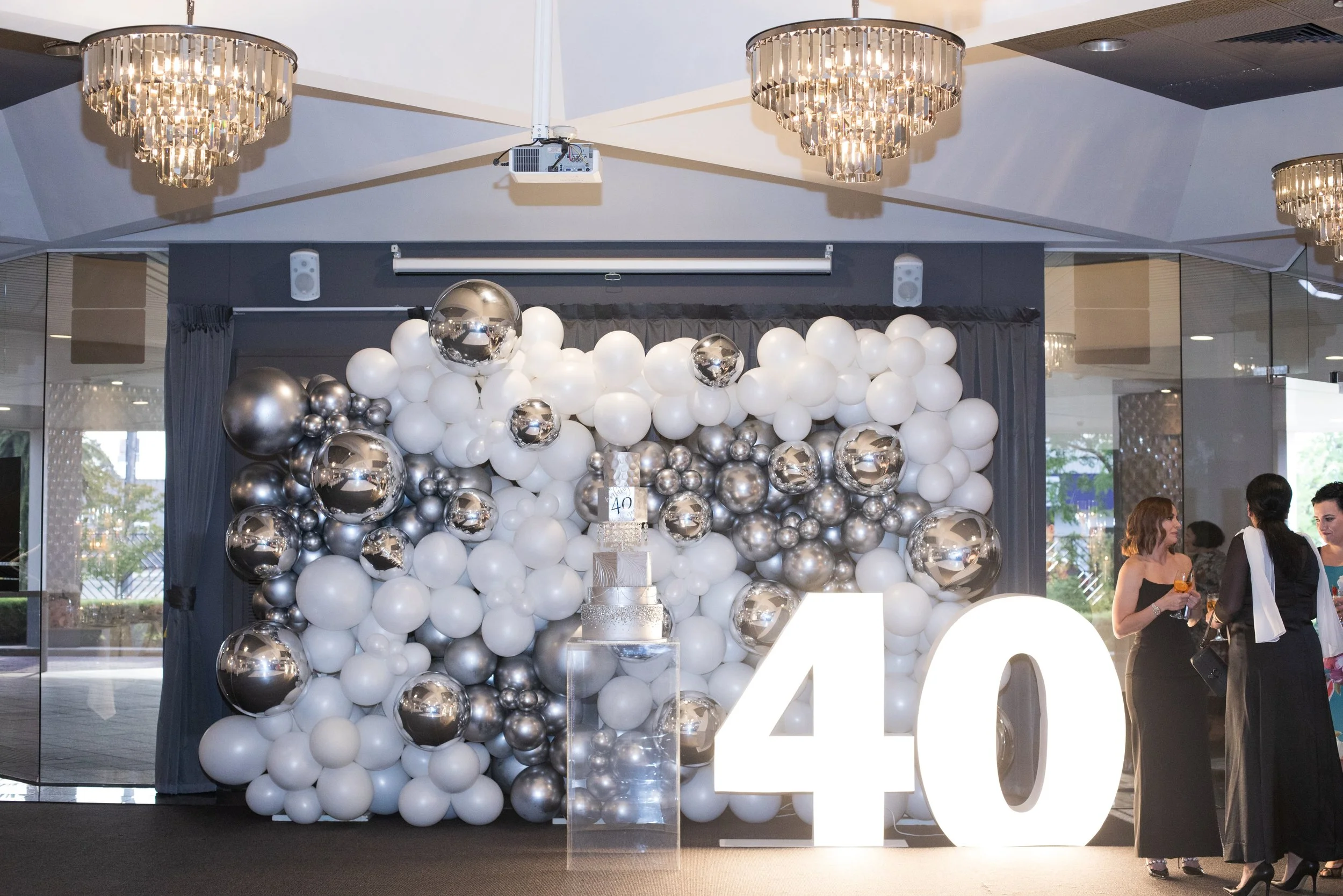 A balloon arrangement featuring white, silver, and metallic balloons, with large white illuminated letters spelling out '40'. There are people in formal attire standing nearby, with chandeliers hanging from the ceiling and a cake displayed in front o