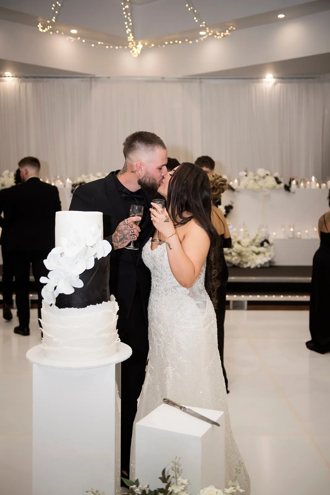The Bride & Groom celebrating with a glass of champagne & kissing each other on white decorated hall.