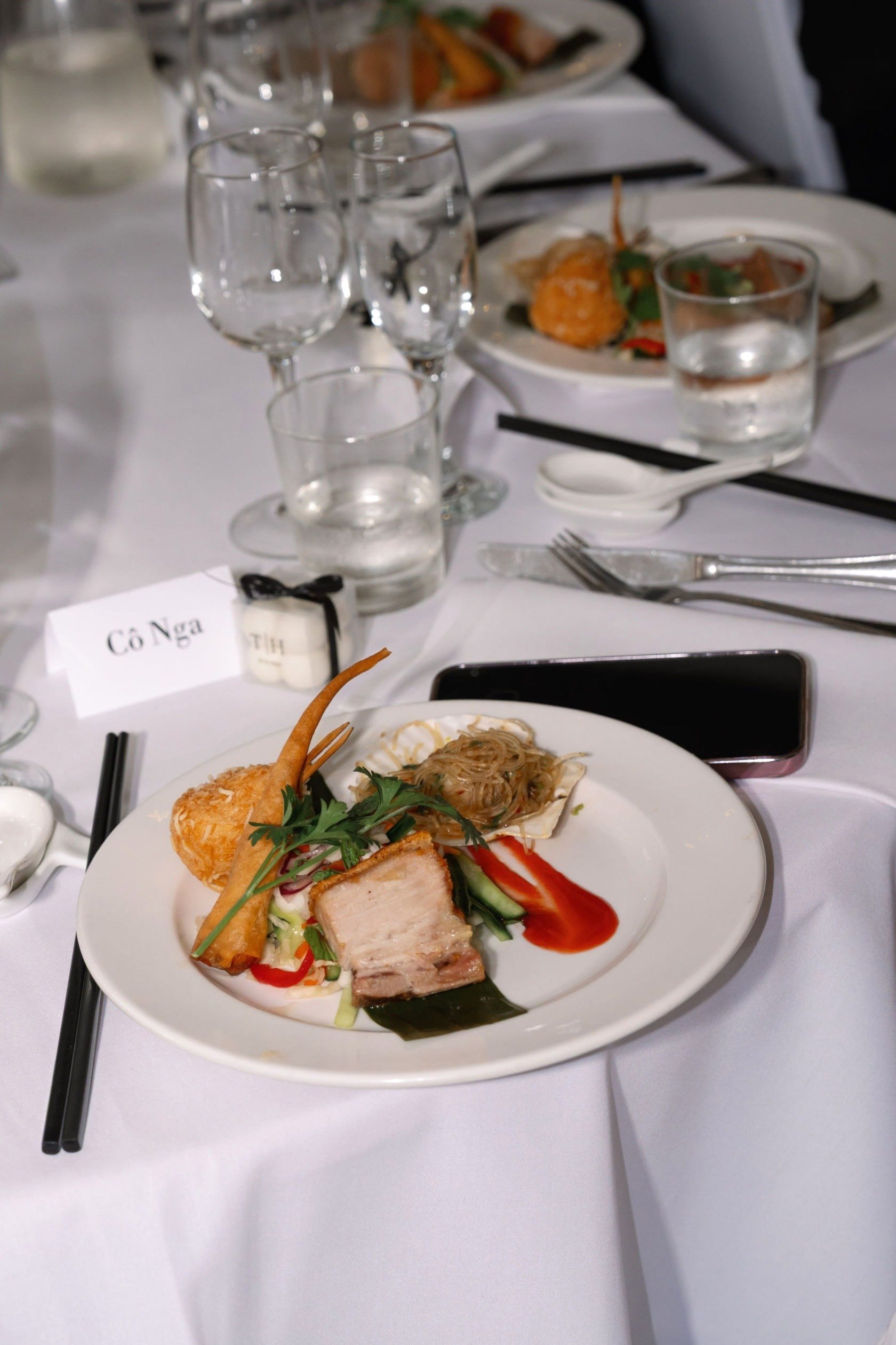 A table with a white tablecloth set at a formal dining event. The focus is on a white plate of assorted foods, including slices of meat, vegetables, noodles, and a breaded item. There are chopsticks, a fork, and a spoon on the table, along with glass