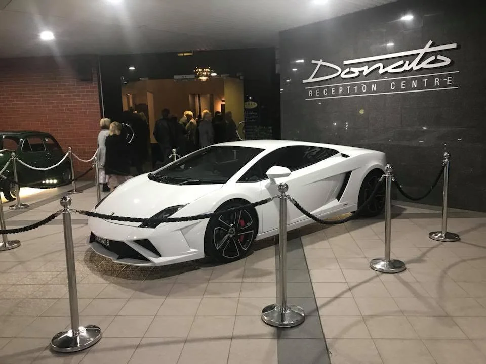 A white Lamborghini sports car displayed inside the Donato Reception Centre, surrounded by black stanchions with ropes. People are seen in the background near the entrance at Donato reception centre in Adelaide