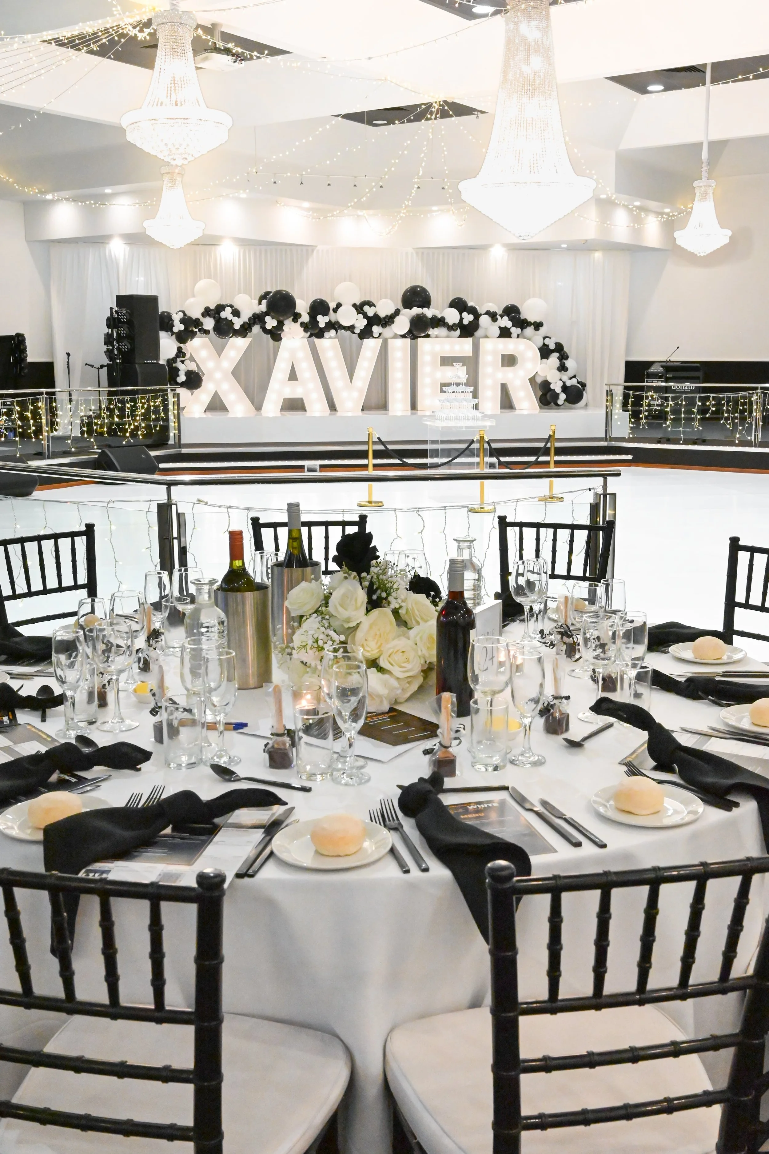 A banquet hall decorated for a celebration with a large sign spelling 'XAVIER' illuminated with lights, surrounded by black and white balloons and fairy lights. There is a dais with chandeliers and a champagne tower in the background, and a large tab