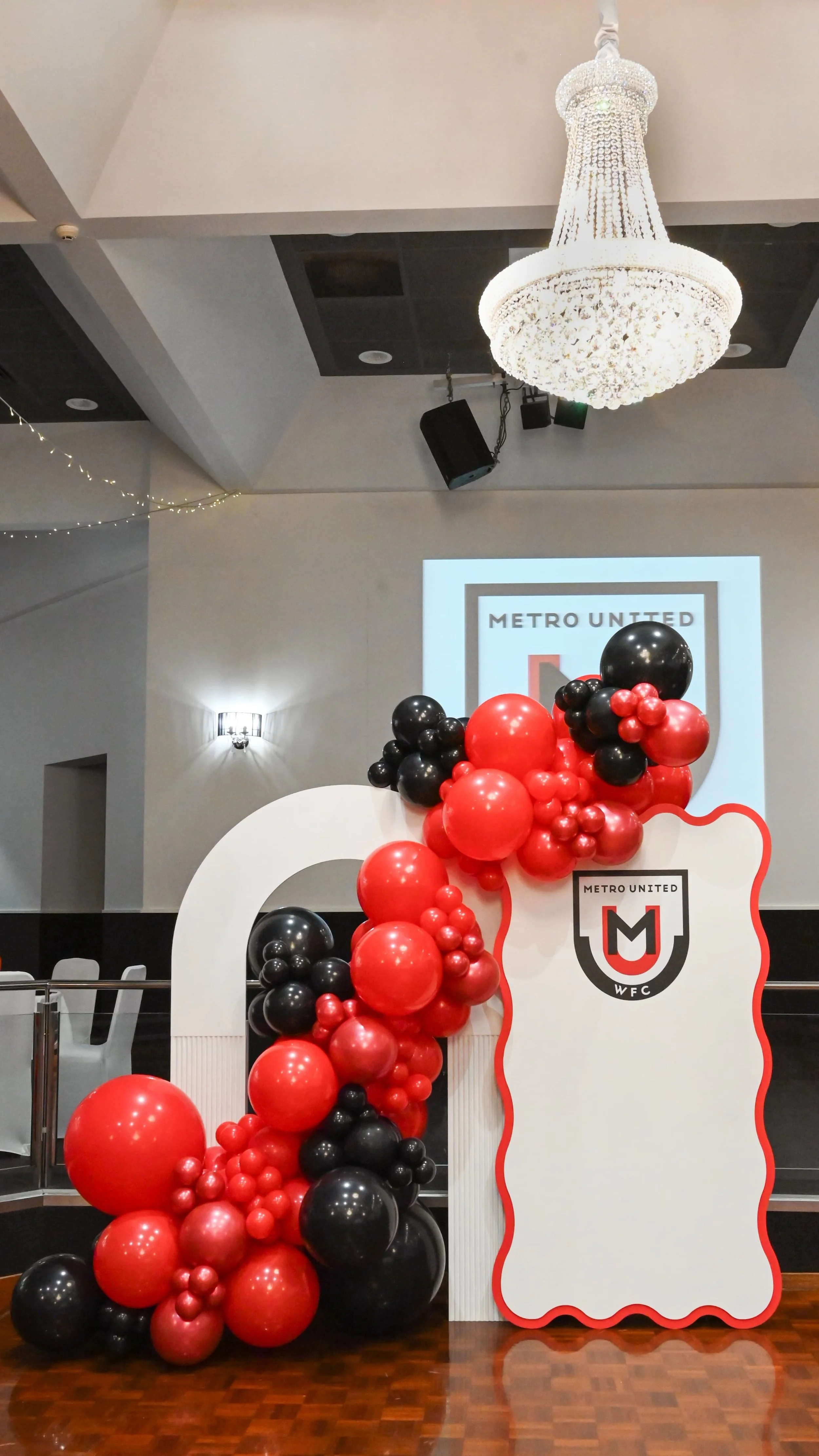 Decorative display with red and black balloons arranged around a white sign with event information, in front of a room with a chandelier and a speaker.