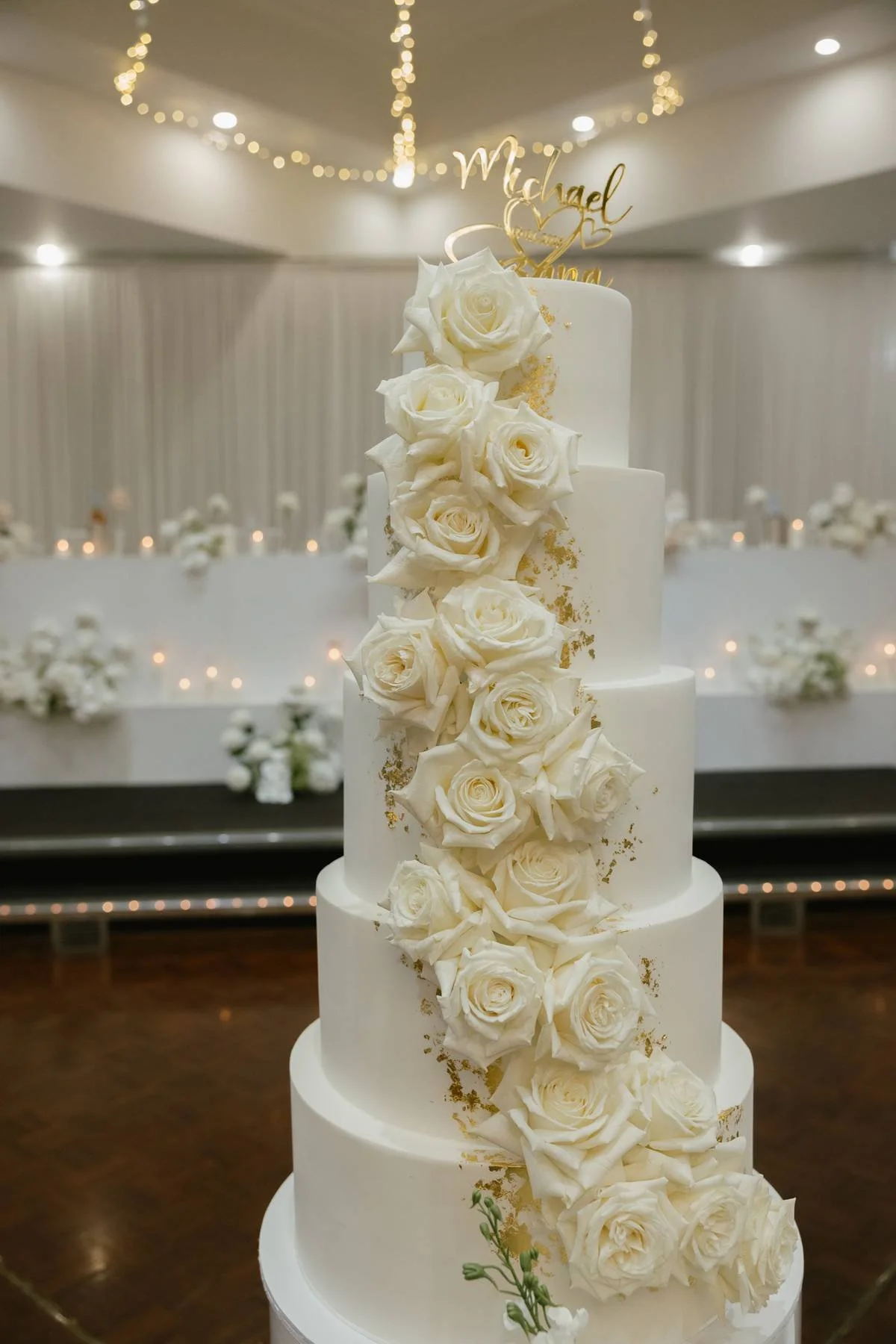 A multi-tiered white wedding cake decorated with cascading white roses and gold accents, topped with a gold cake topper that reads 'Michael' with hearts.