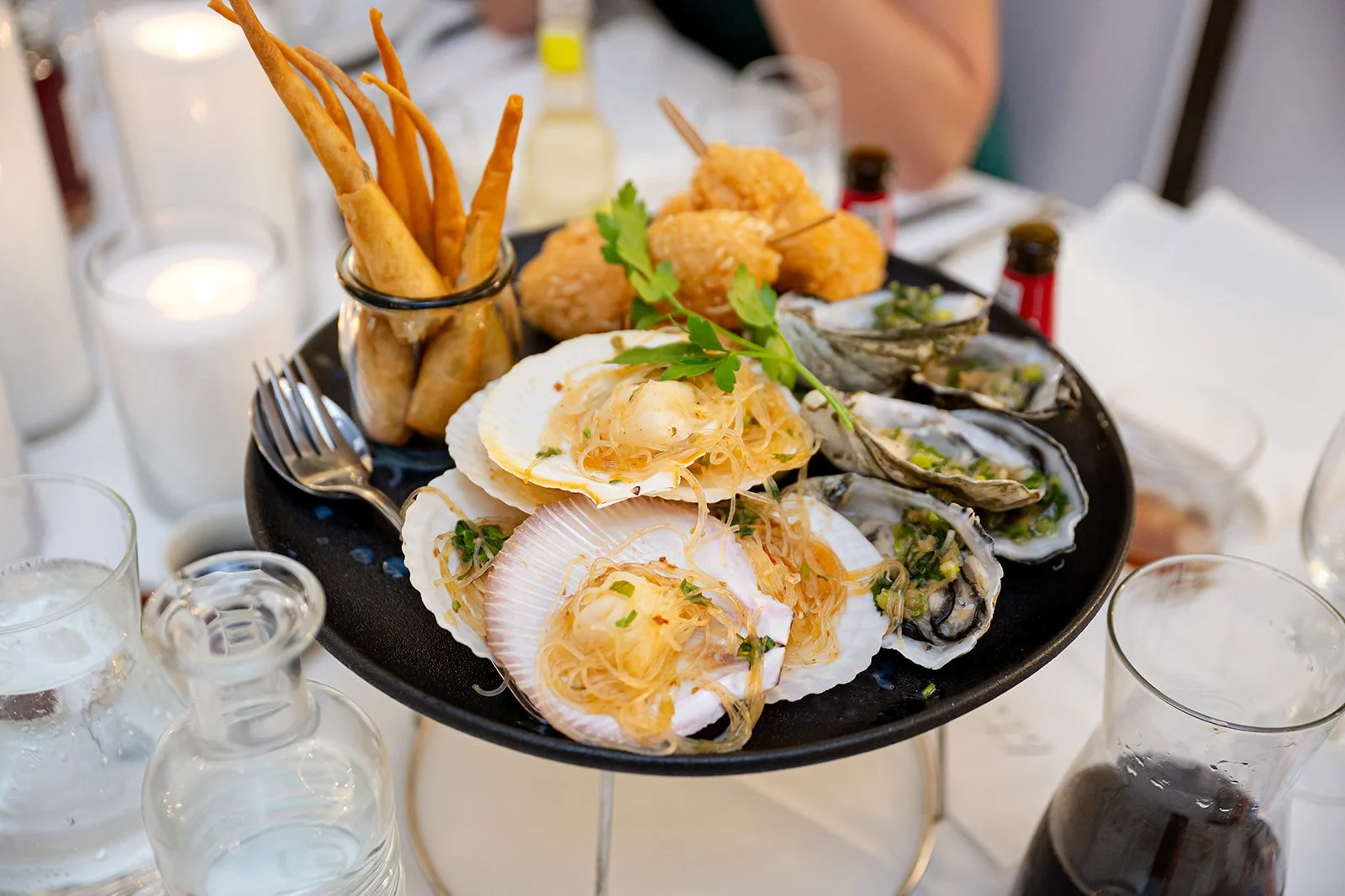 A black tray with assorted seafood including scallops in shells topped with sauce and herbs, oysters with green onion and sauce, fried chicken bites with a sprig of parsley, and crispy breadsticks in a glass container, set on a table with glasses and