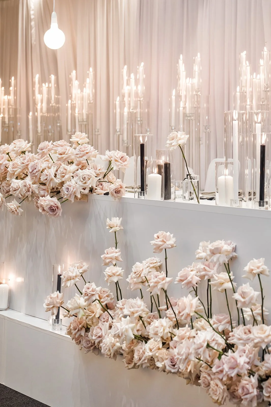 Elegant wedding or event decor with pastel pink roses, white candles, and tall candle holders on a white table, with sheer white curtains and warm lighting in the background.