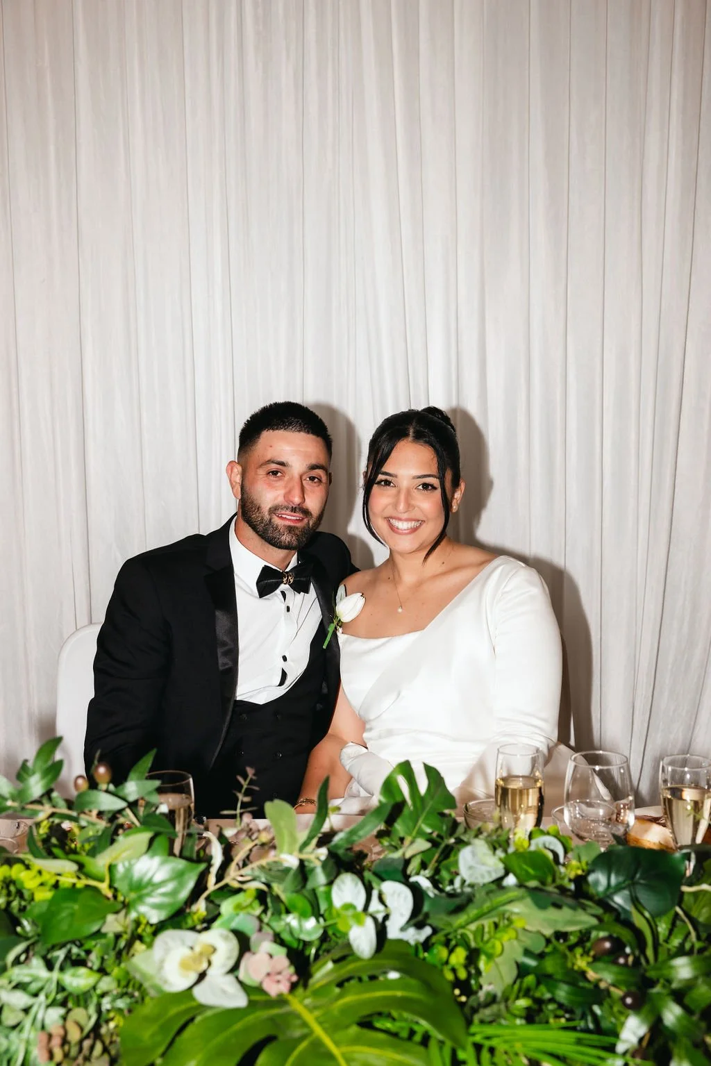 A bride and groom sitting together at their wedding reception, smiling, with a white background and a floral centerpiece on the table at Donato reception centre in Adelaide
