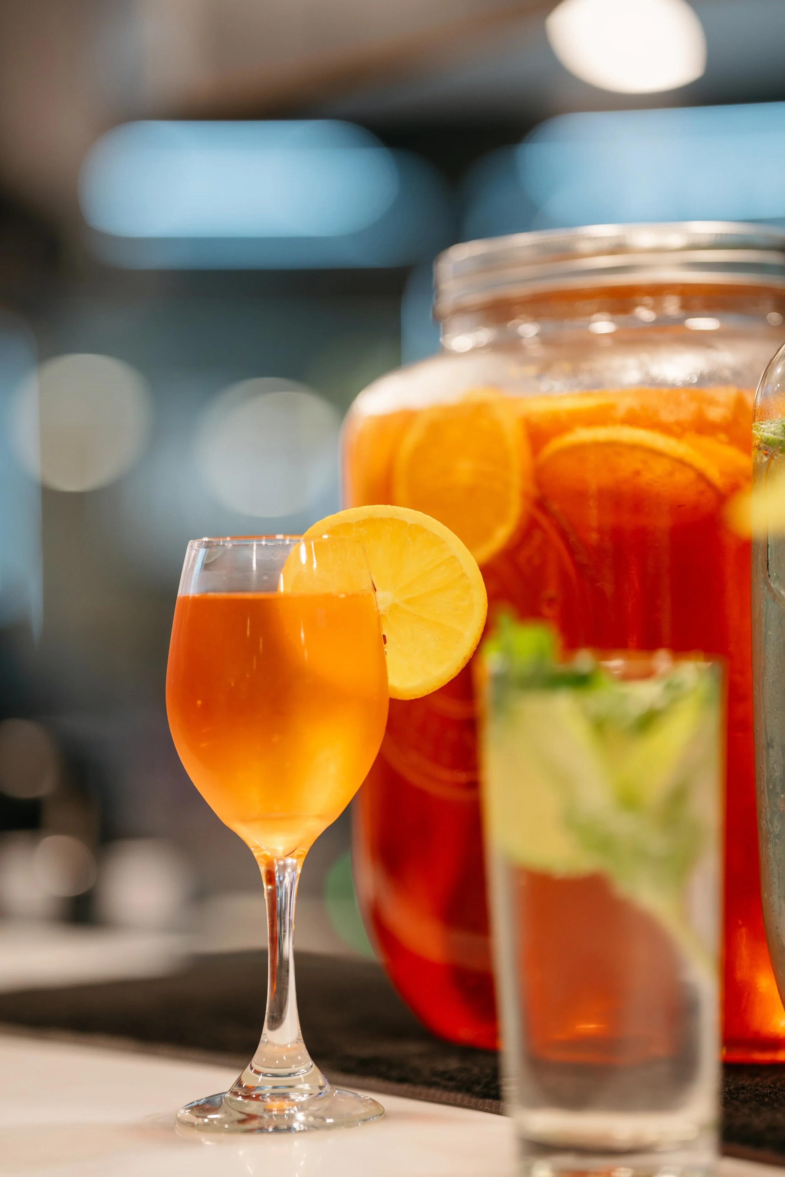 A glass of orange-colored beverage garnished with a lemon slice in the foreground, with a jar filled with sliced oranges and a pitcher of the same drink in the background.