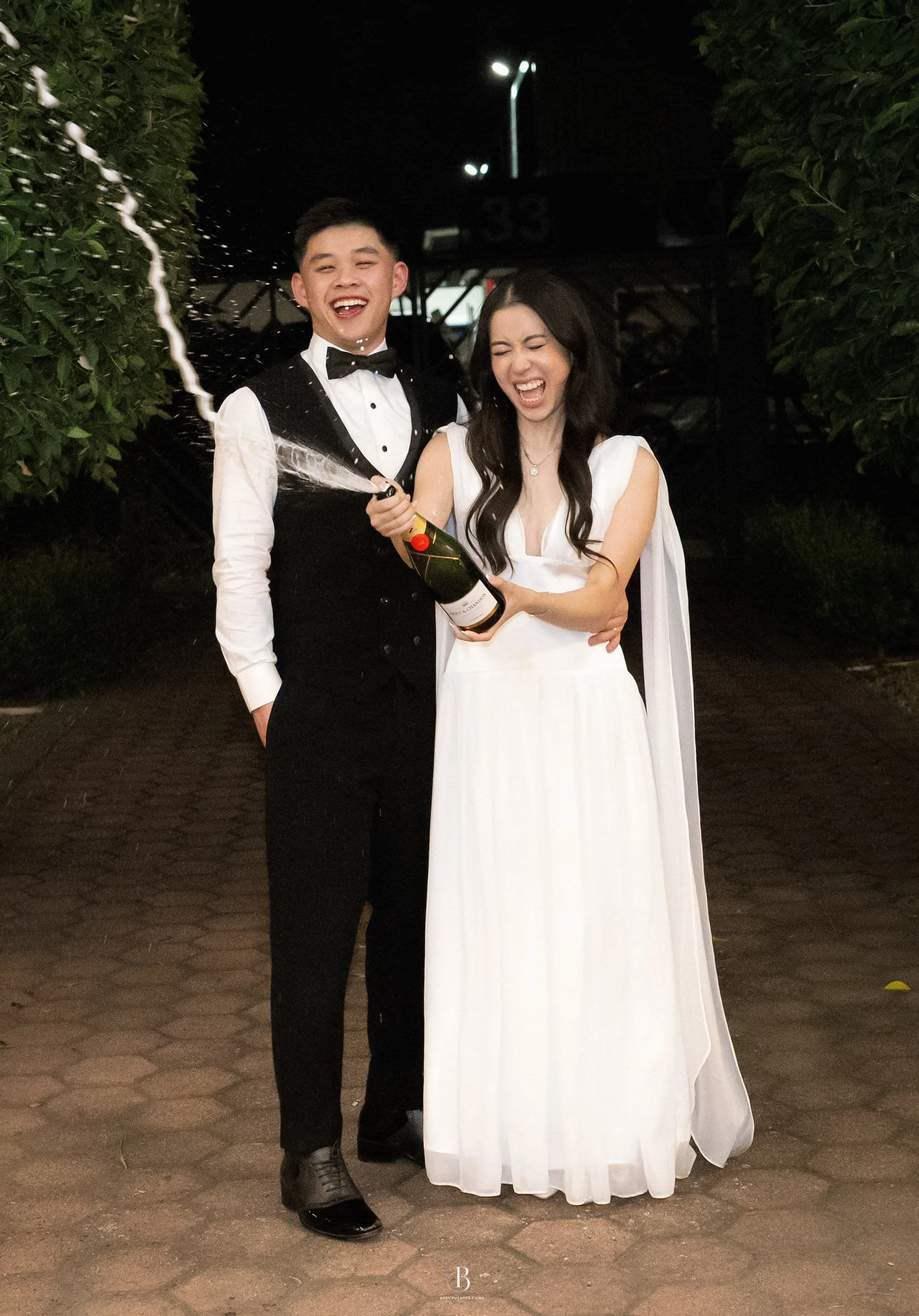 A young man in a tuxedo and a woman in a white dress celebrating with a bottle of champagne outdoors at night, with trees and streetlights in the background at Donato reception centre in Adelaide