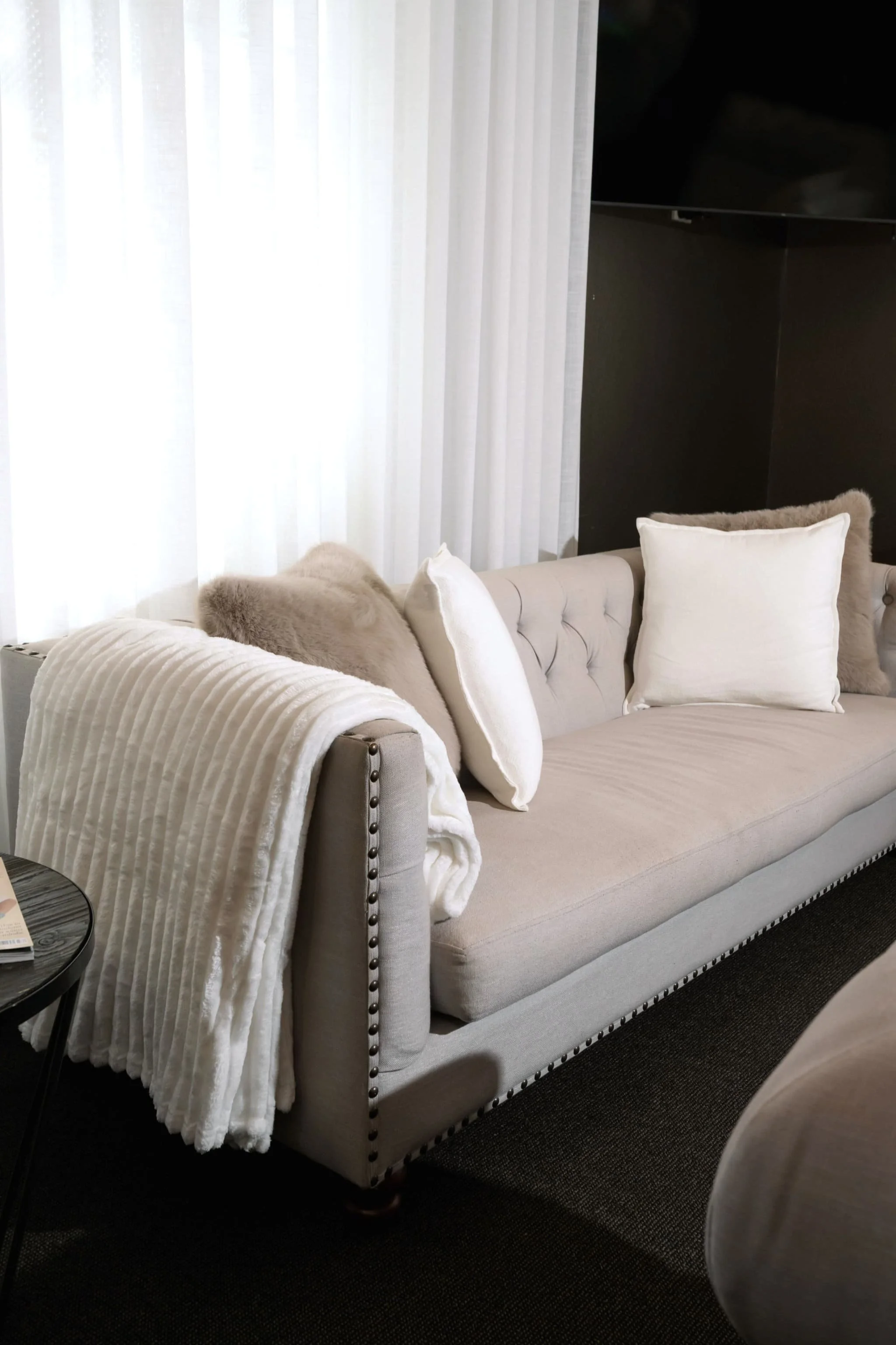 A light-colored sofa with rolled arms and nailhead trim, decorated with white and beige pillows and a faux fur throw blanket, located next to a small black side table with a magazine on it. Behind the sofa, there are sheer white curtains covering a w