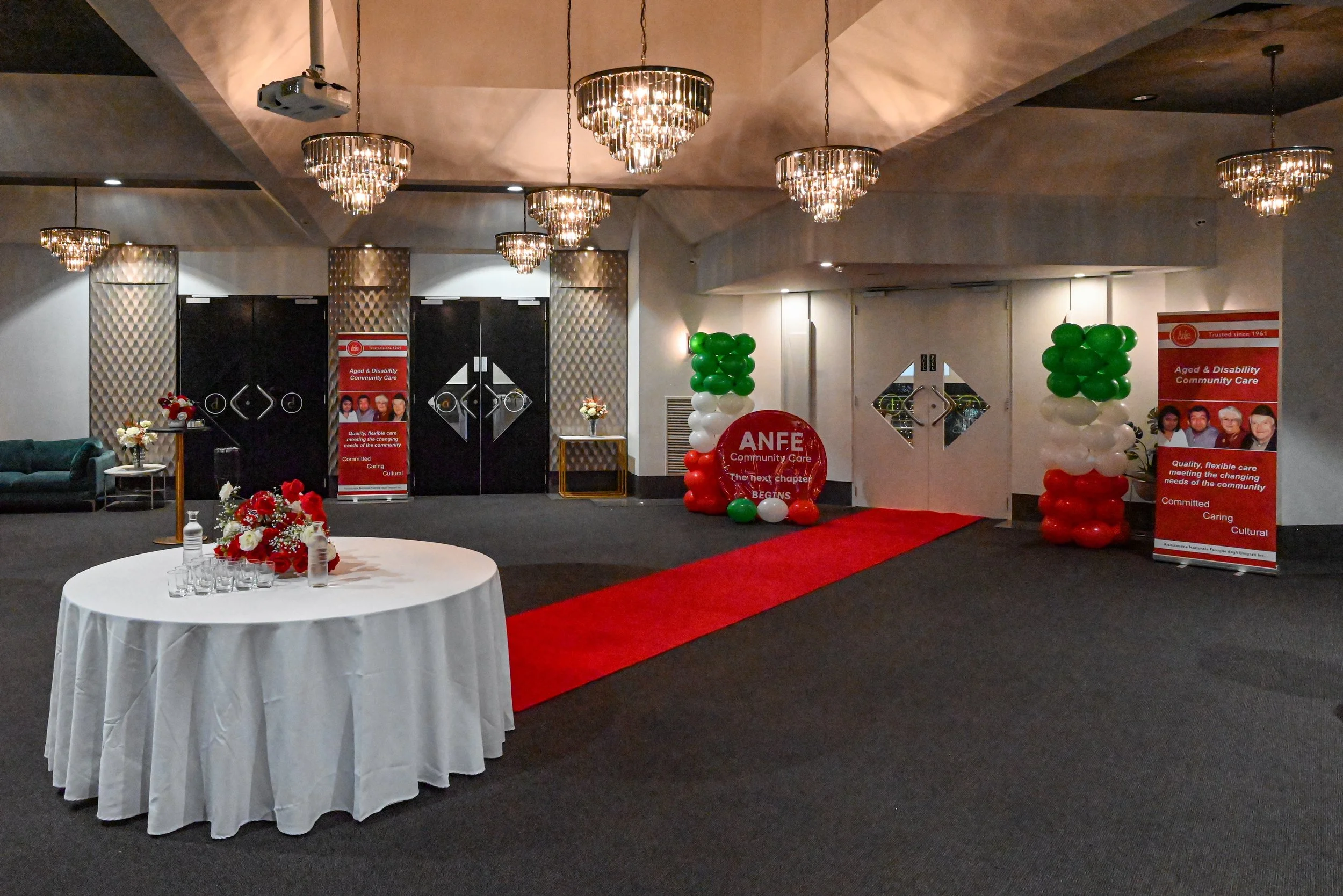 Event hall decorated with banners and balloons in red, white, and green for an organization named ANFE Community Care, featuring chandeliers hanging from the ceiling, a table with flowers, and a red carpet.