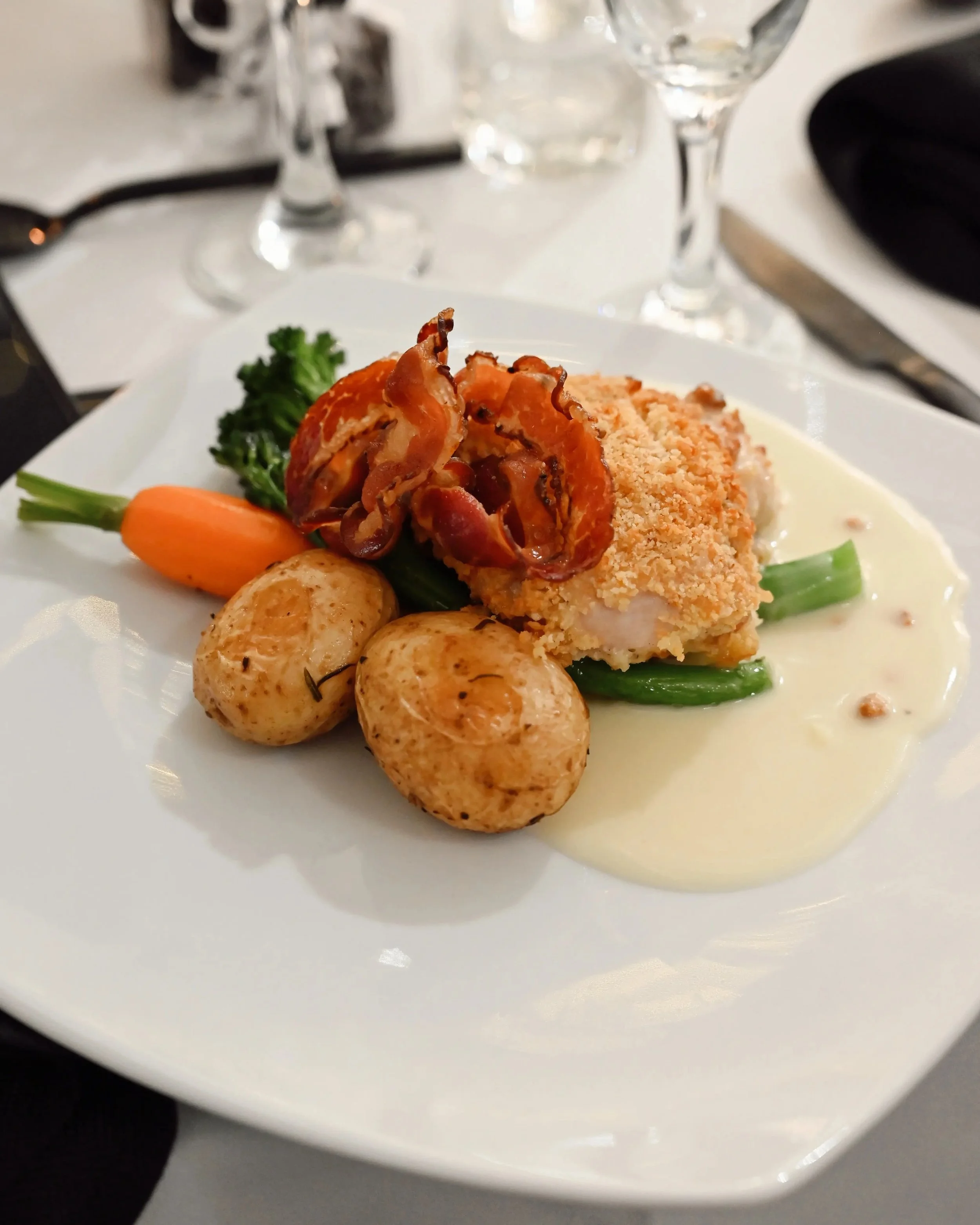 Plate of food including a piece of baked fish topped with crispy bacon, roasted potatoes, broccoli, and a carrot, served with a creamy sauce