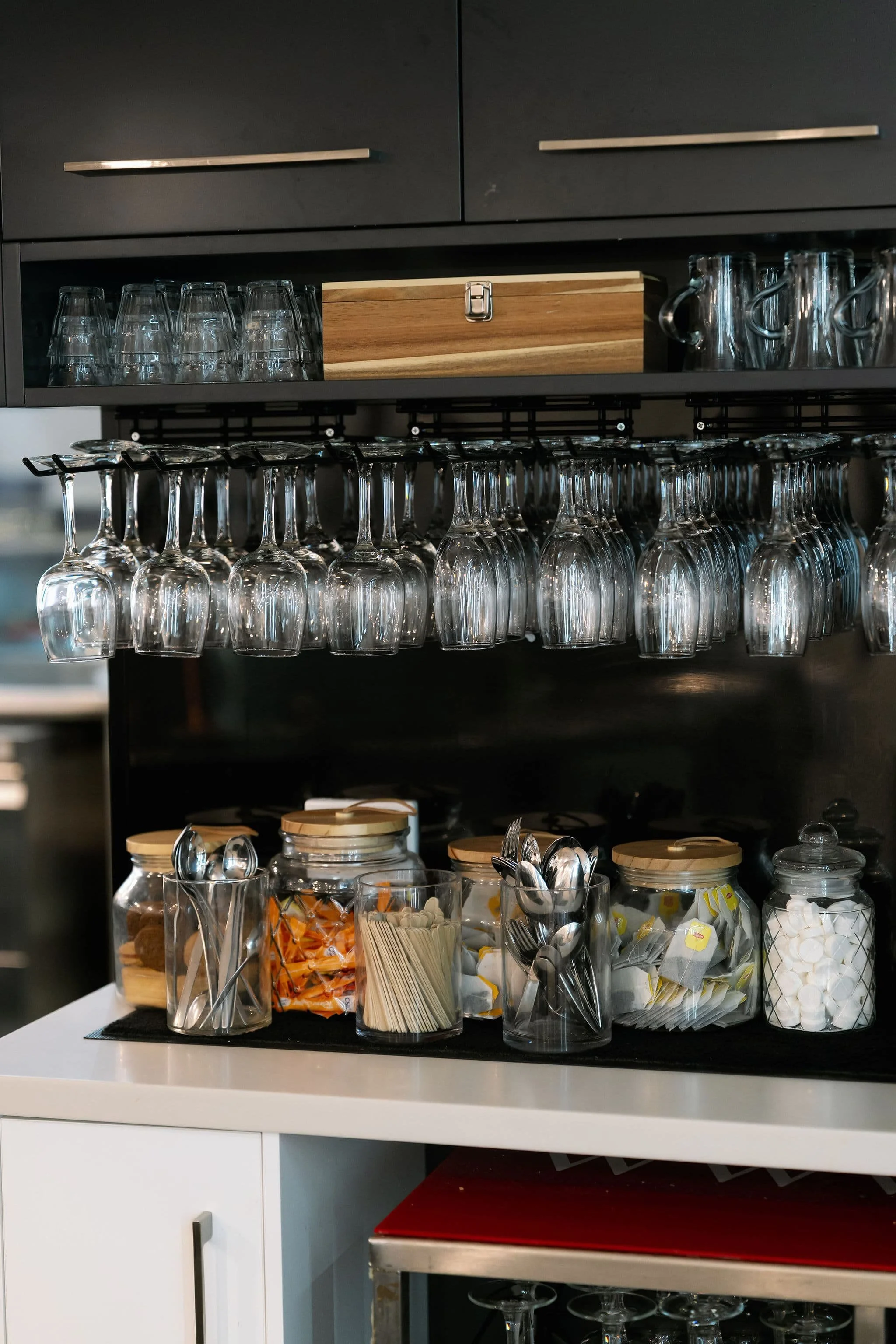Kitchen shelf with upside-down wine glasses, a wooden box, jars of snacks, plastic utensils, and sugar packets on a white countertop.