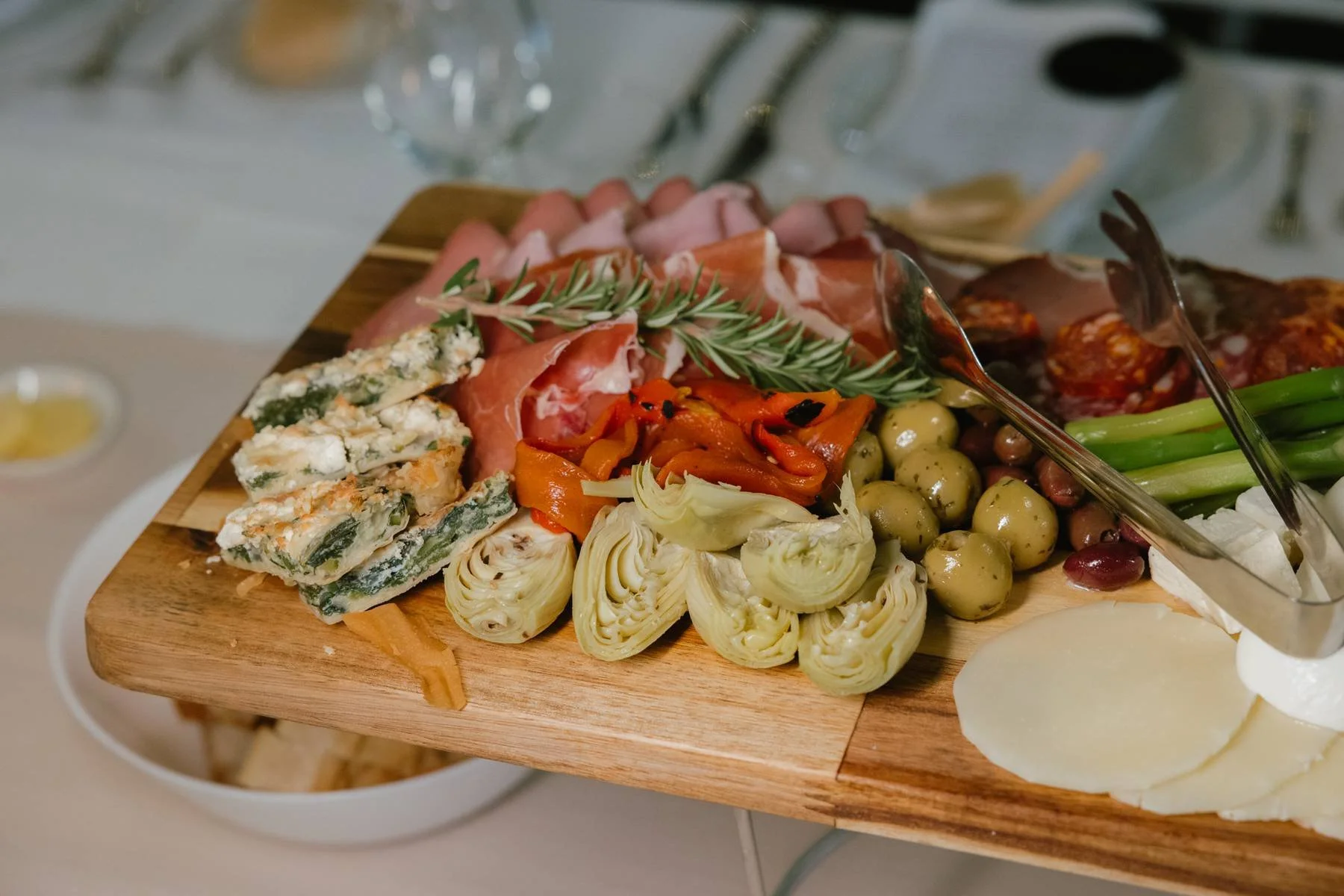 A wooden serving board with assorted antipasto including cured meats, artichoke hearts, mixed pickled vegetables, olives, blue cheese, sliced tomatoes, green onions, and fresh herbs.