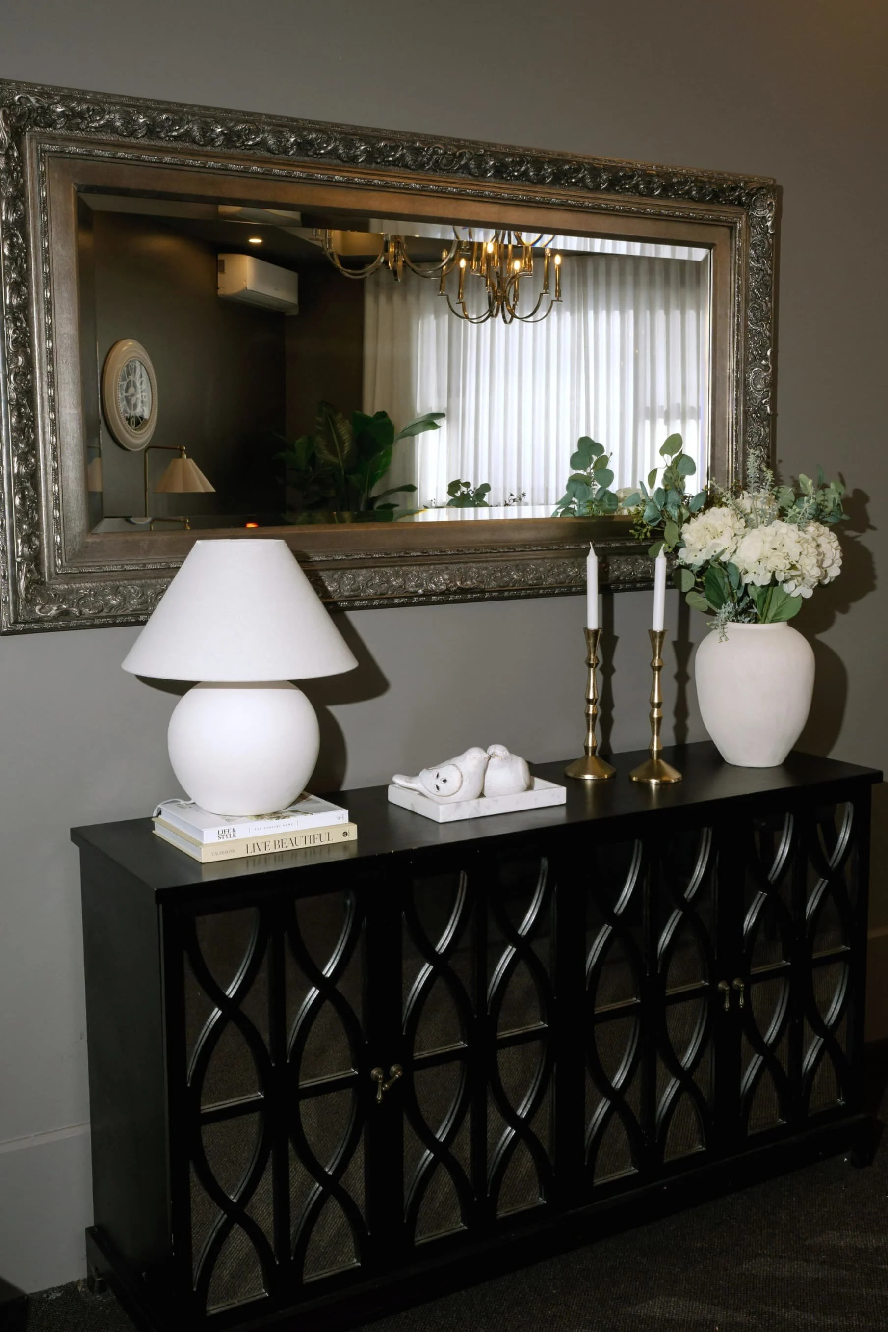 A black console table with an intricate circular pattern on the front, topped with a white lamp, a white vase with white flowers and green leaves, two gold candlesticks with white candles, and a small white decorative sculpture. Behind, there is a mi