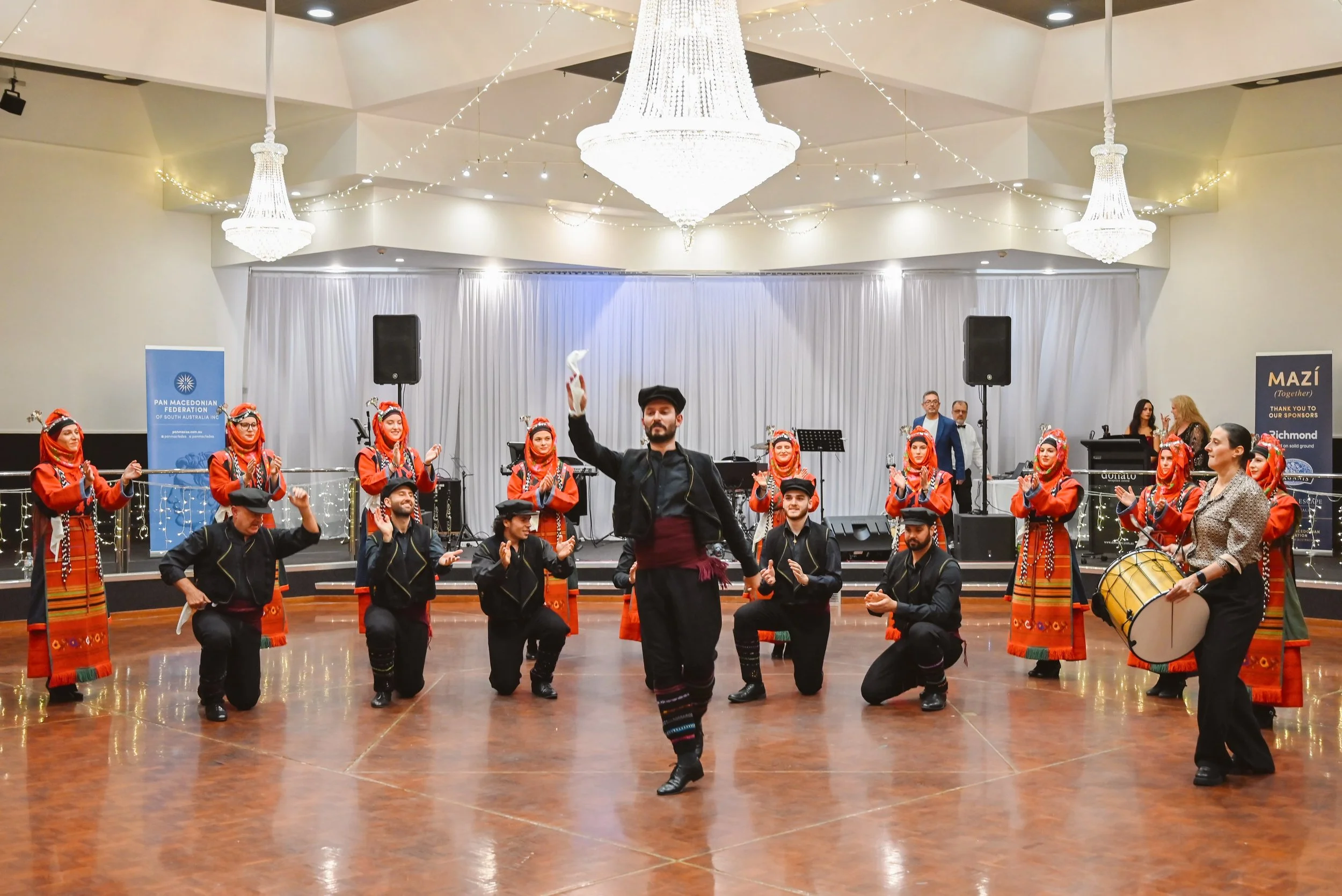 Group of performers in traditional colorful clothing dancing on a wooden floor, with a woman playing a drum and men holding a hand-held drum, in a decorated indoor venue with chandeliers and string lights.