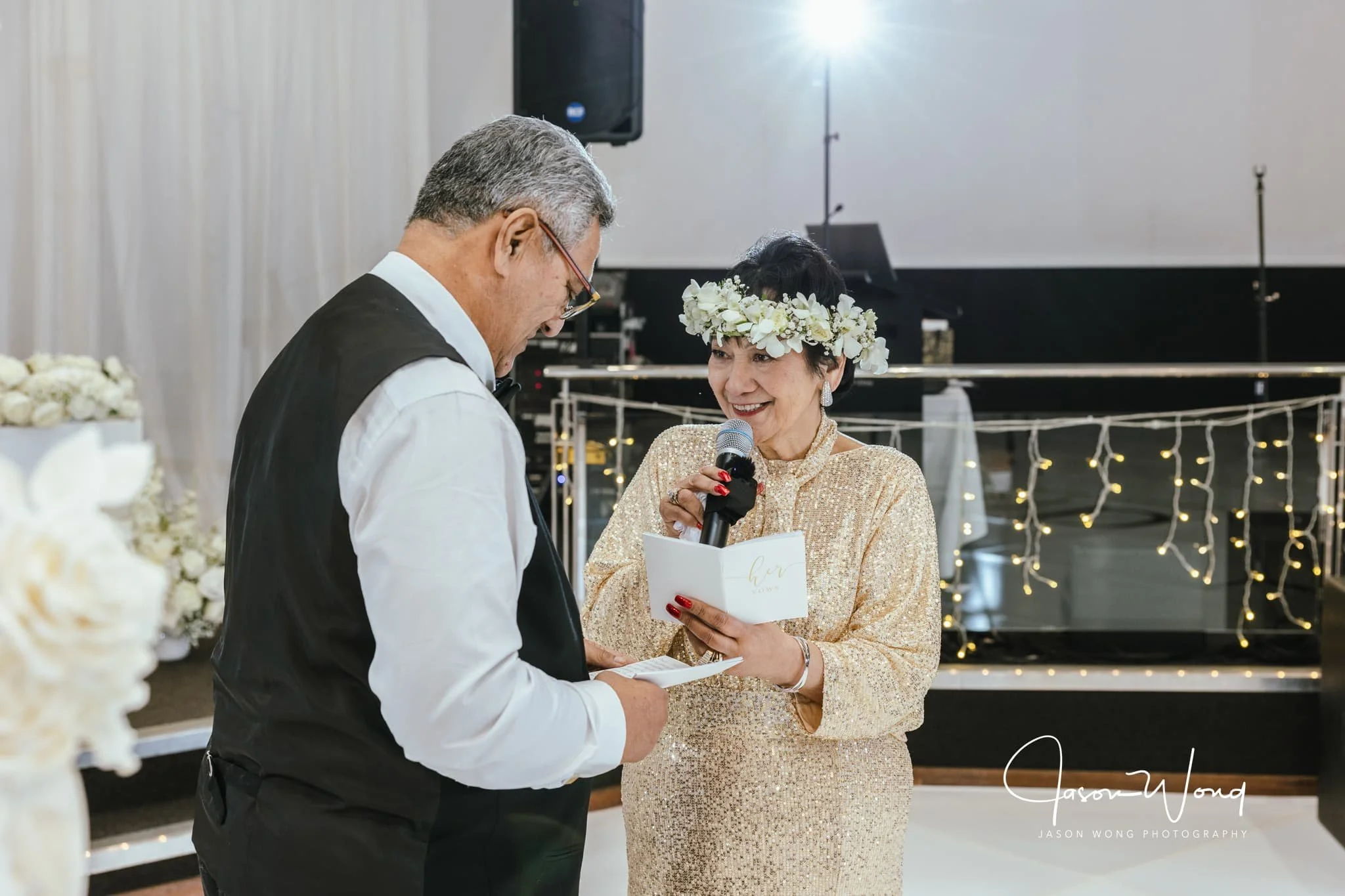 A woman wearing a floral crown and a gold sequined dress is reading vows into a microphone while a man dressed as a groom holds her hands during a wedding ceremony, with floral decorations and fairy lights in the background.