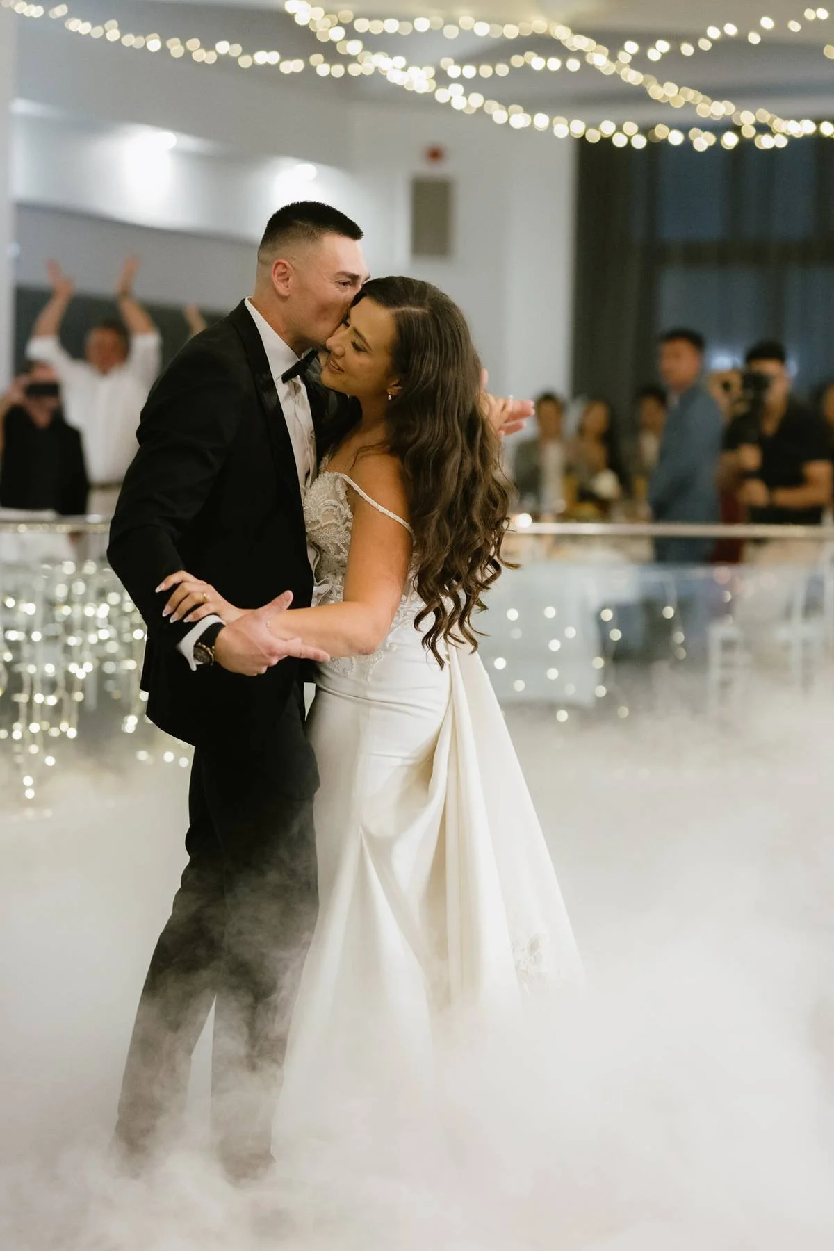 A bride and groom dancing at a wedding reception, surrounded by friends and family. The bride is wearing a white gown with lace details, and the groom is in a black tuxedo. The background has string lights and guests watching at Donato reception cent
