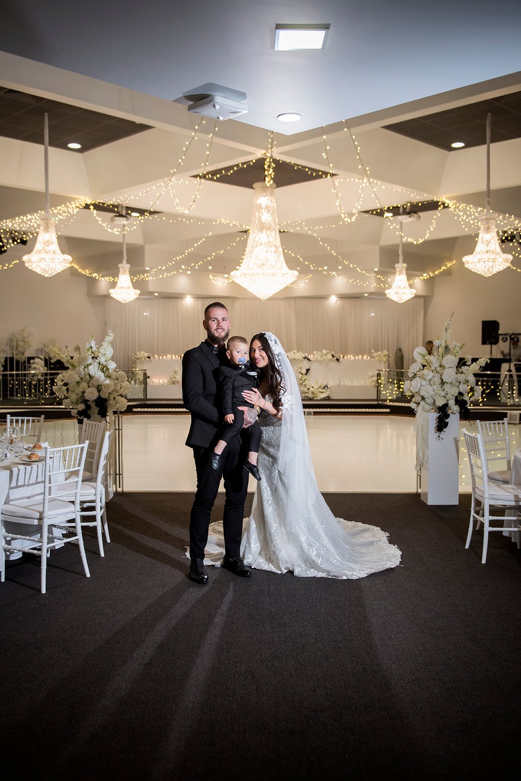 A bride and groom with a young boy at a wedding reception, standing in a decorated banquet hall with chandeliers, string lights, and floral arrangements at Donato reception centre in Adelaide