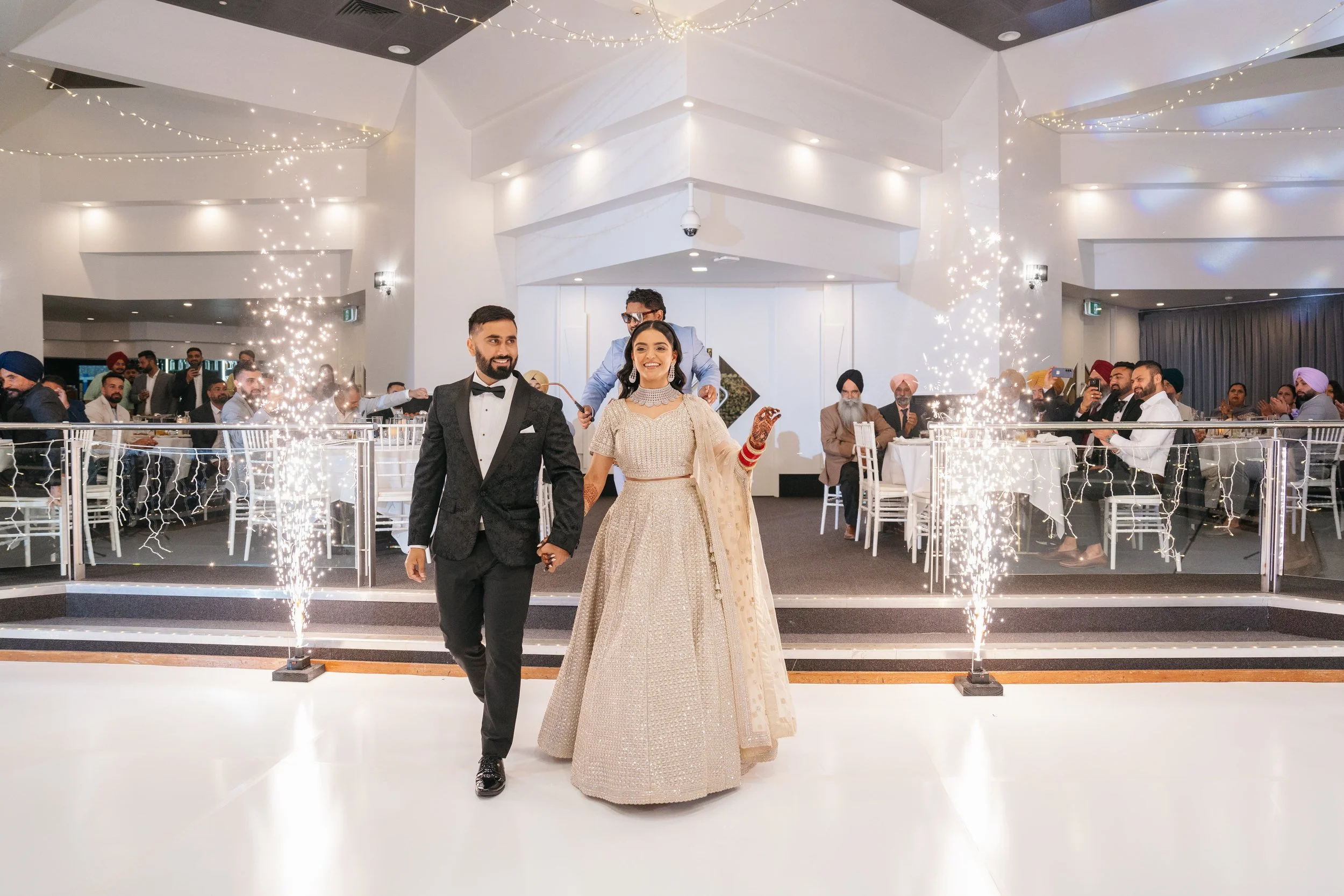 A couple dressed in formal attire, holding hands, walking on a platform with sparklers on either side, at a wedding reception with seated guests cheering in the background.