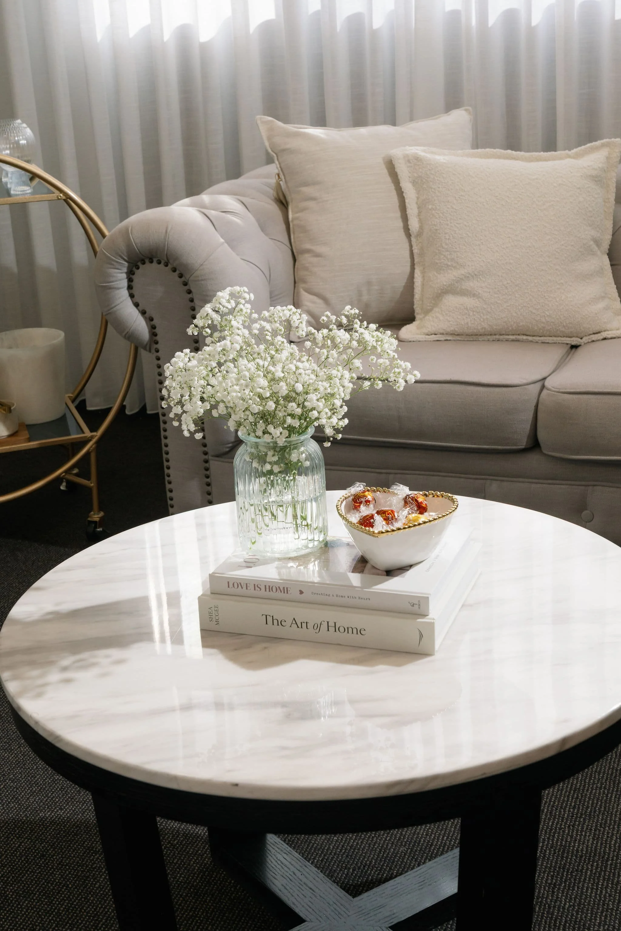 A cozy living room scene featuring a white tufted sofa with beige pillows, a round white marble coffee table with a glass vase of white baby's breath flowers, and books titled 'Love is Home' and 'The Art of Home' on the table. A side table with a gla