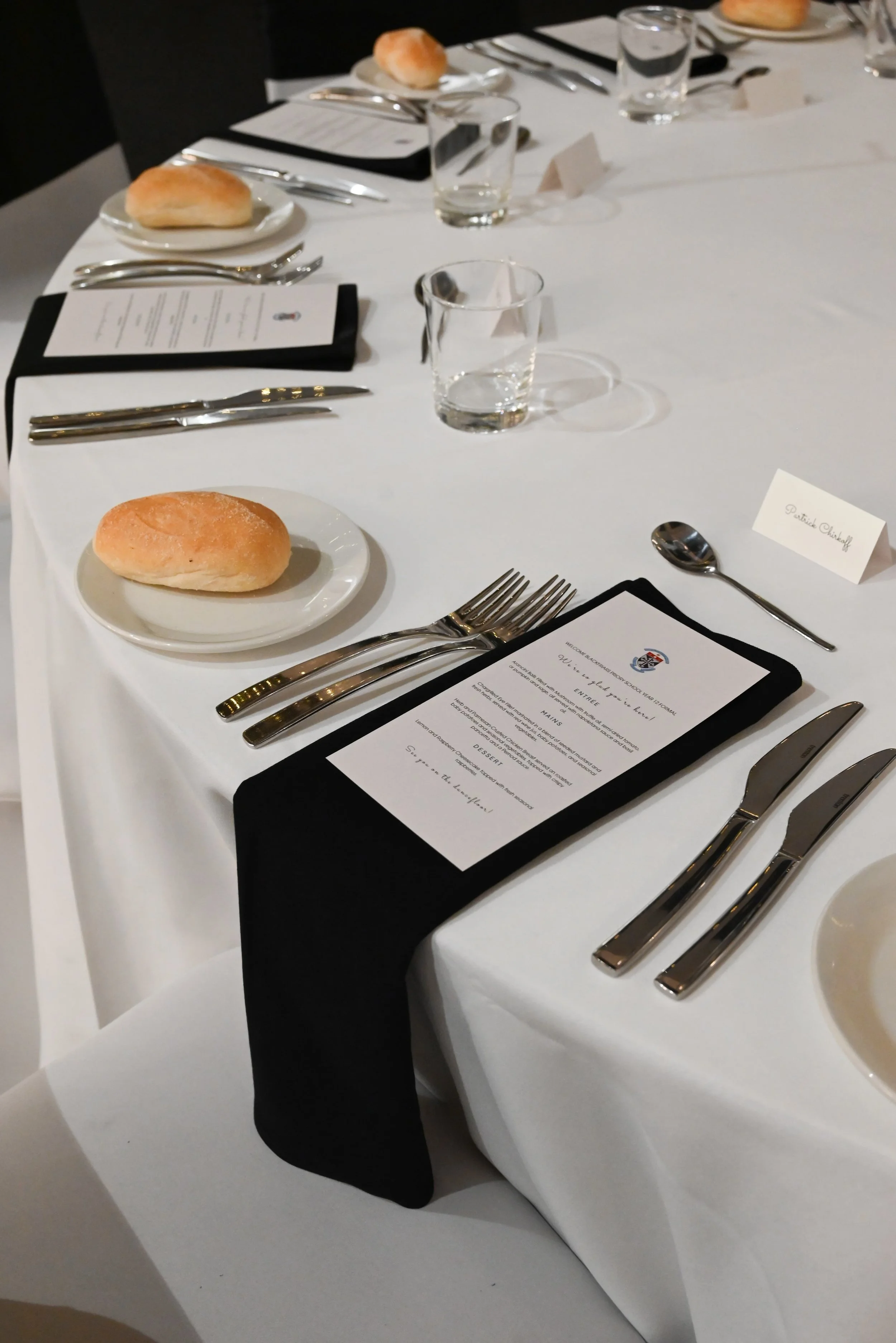 A formally set dining table with a white tablecloth, multiple utensils, clear drinking glasses, bread rolls on small plates, and menus with some place cards, including one with the name 'Patrick Christoff'.