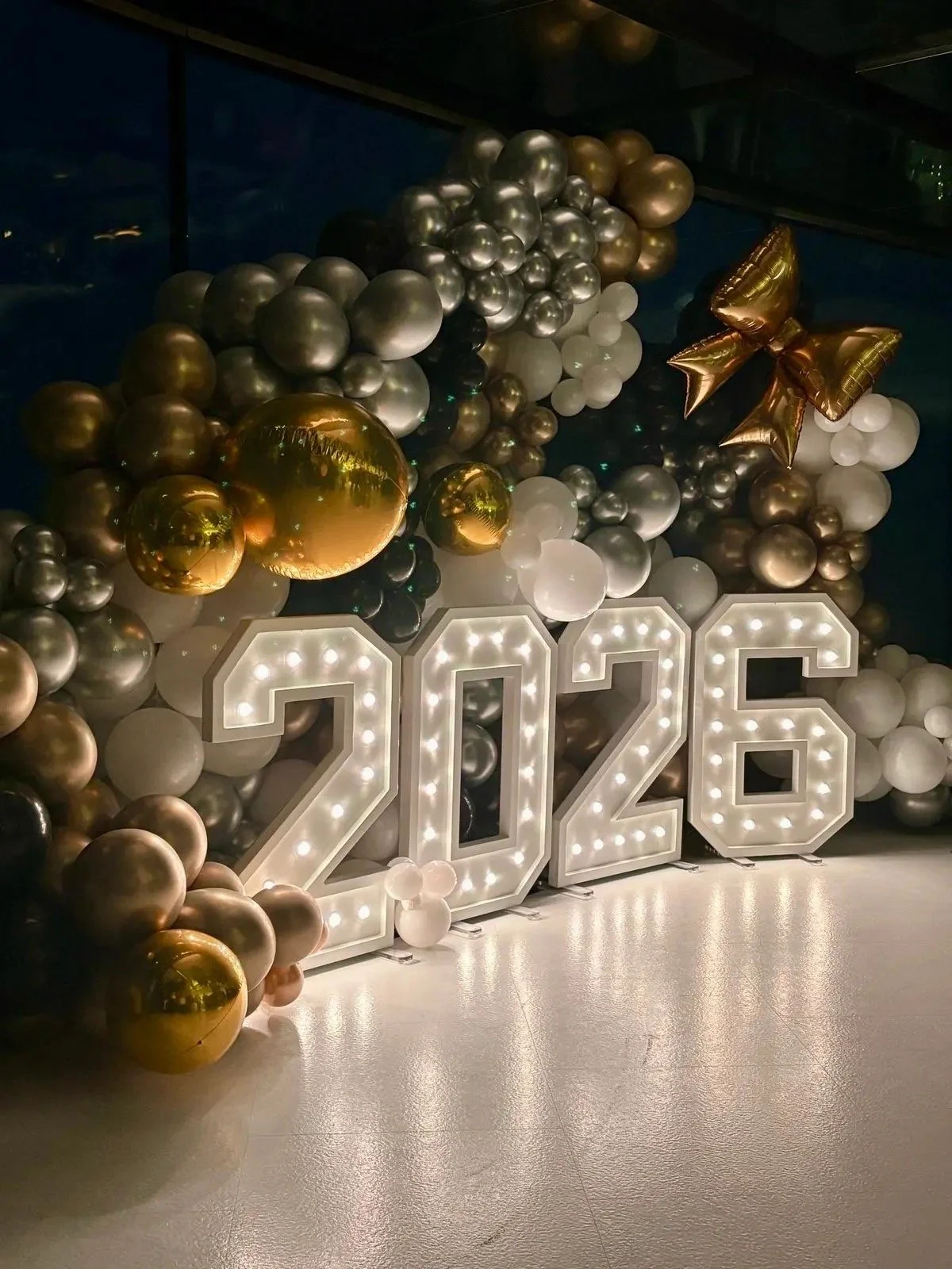 Decorative display featuring large illuminated '2026' numbers surrounded by gold, silver, black, and white balloons, with a large gold bow on the right side.