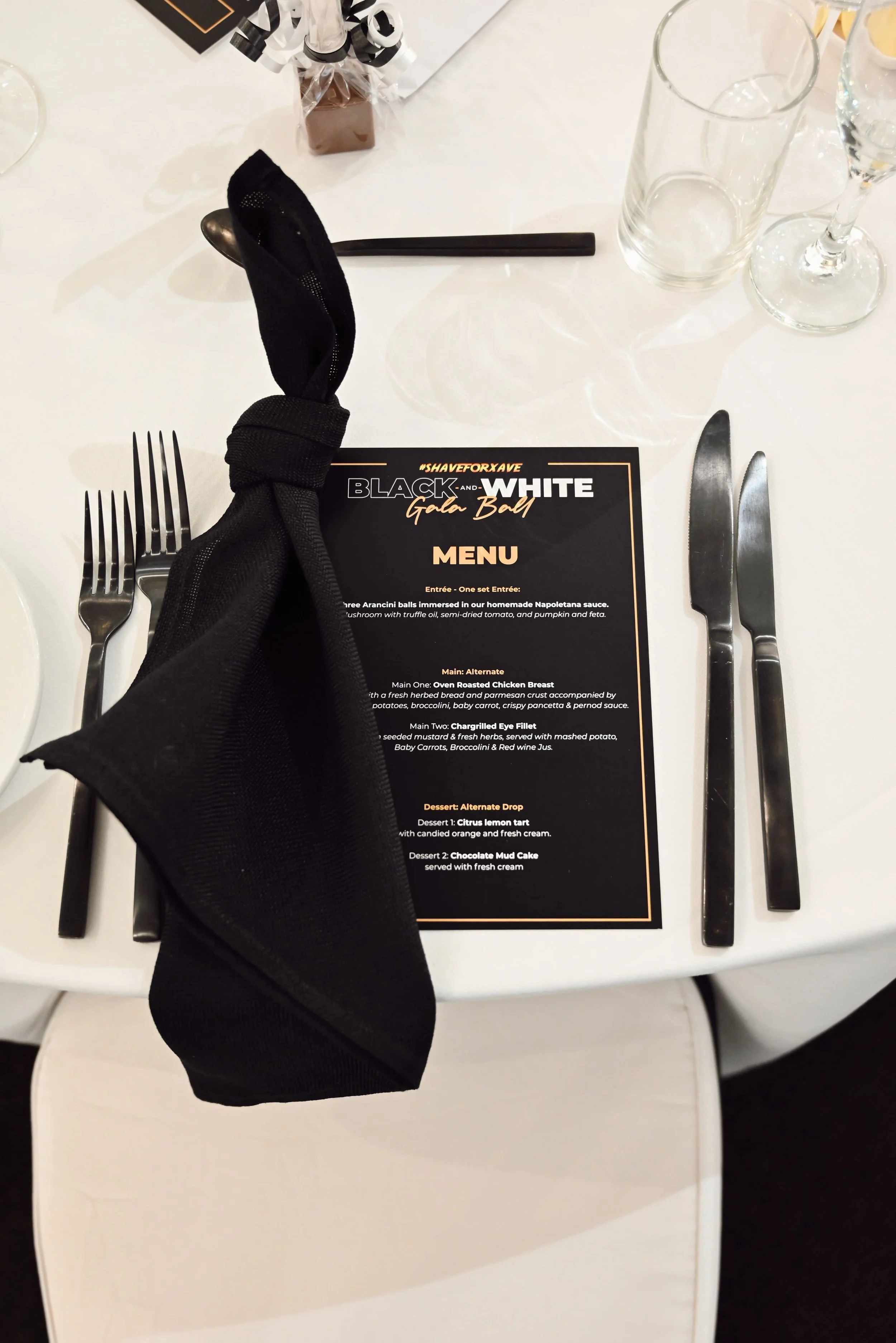 Table setting with black and white event menu, black cloth napkin, silverware on either side of the menu, and empty glasses. The menu features a black background with white and gold text for a formal event.